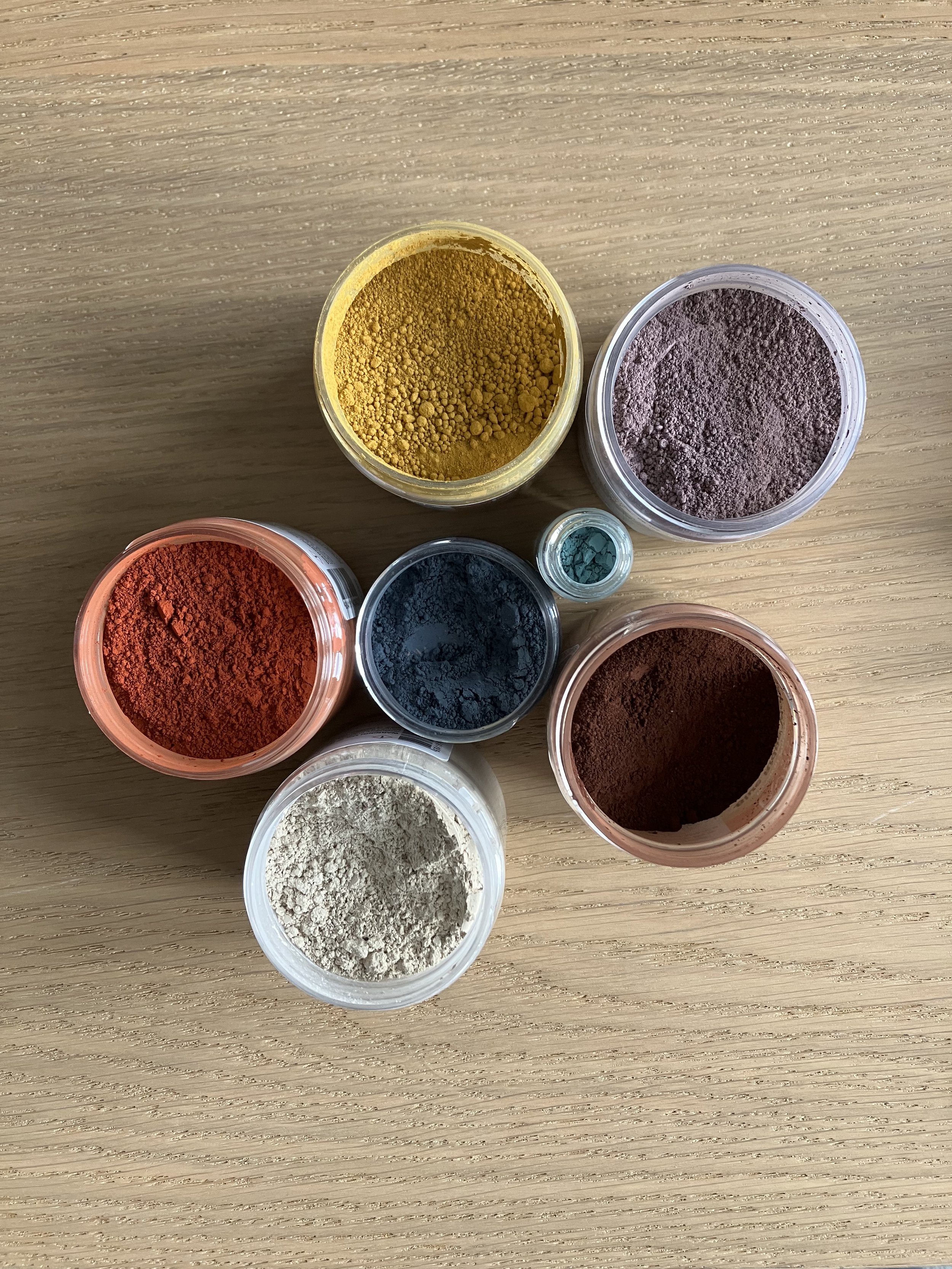 Multiple containers of powdered earth pigments in various colors including yellow, purple, red, black, white, and brown, placed on a wooden surface.
