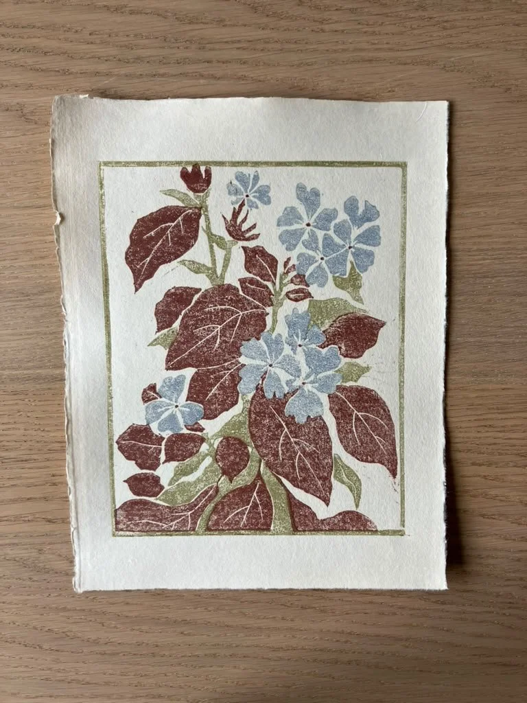 A piece of paper with a floral illustration featuring brown and green leaves and light blue flowers, placed on a wooden surface.