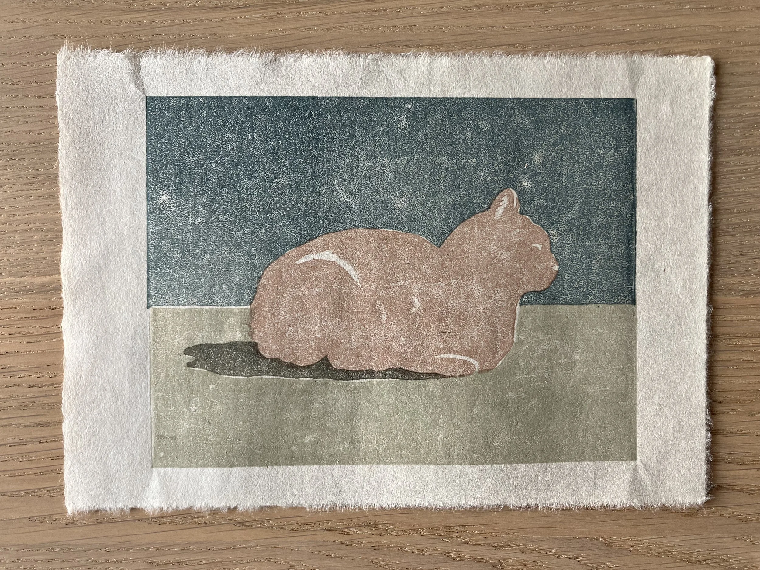 A woodcut depicting a loafing orange cat, with green ground and blue sky.