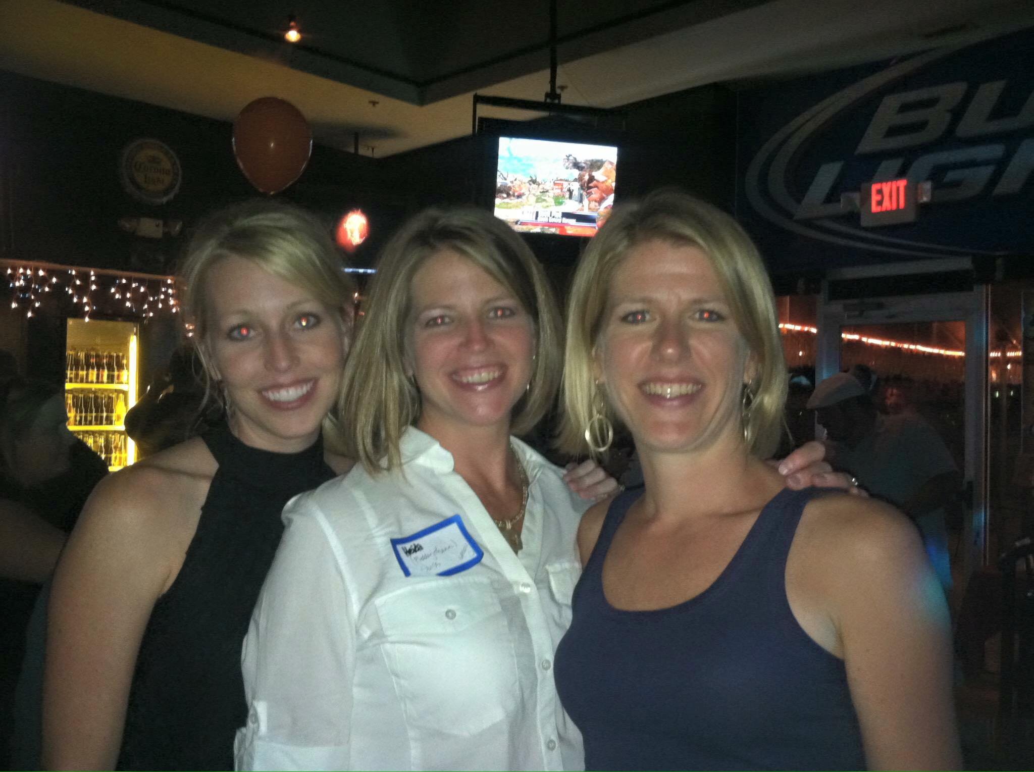 Three women smiling in a dimly lit indoor setting, with artwork and a television screen on the wall in the background.