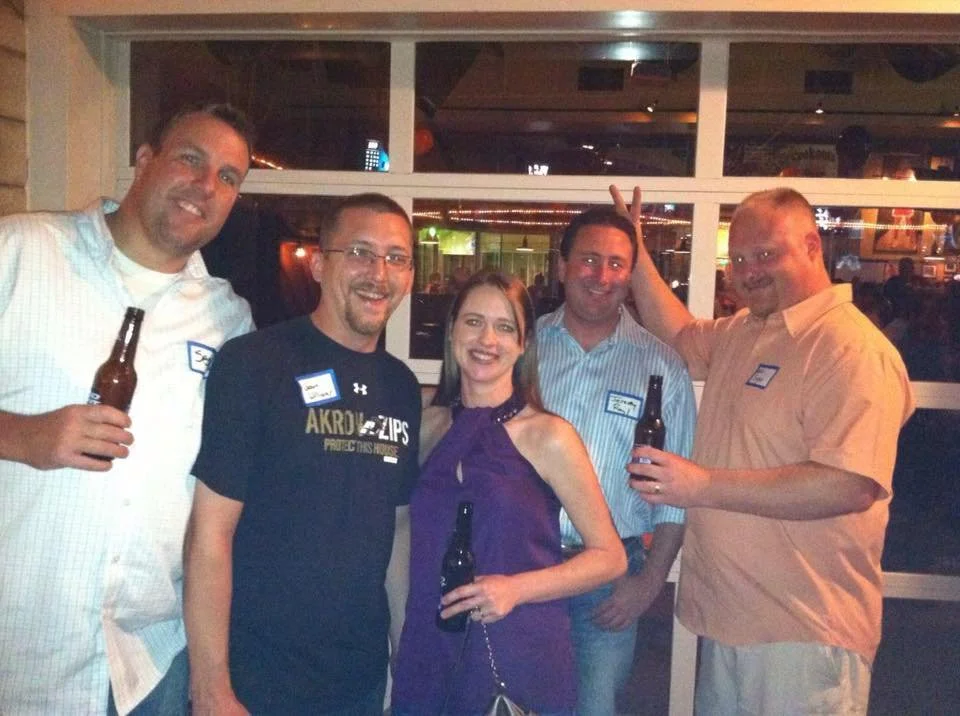 A group of five people, four men and one woman, standing indoors at a social gathering, holding beer bottles and smiling at the camera.