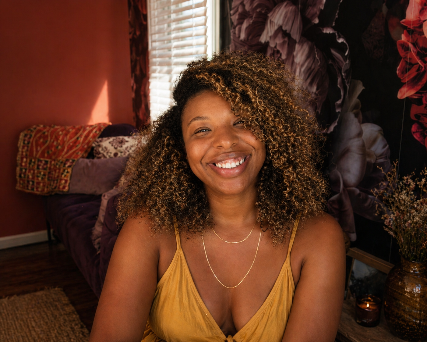 A woman with curly, light brown hair and a bright smile sitting in a cozy, decorated room.