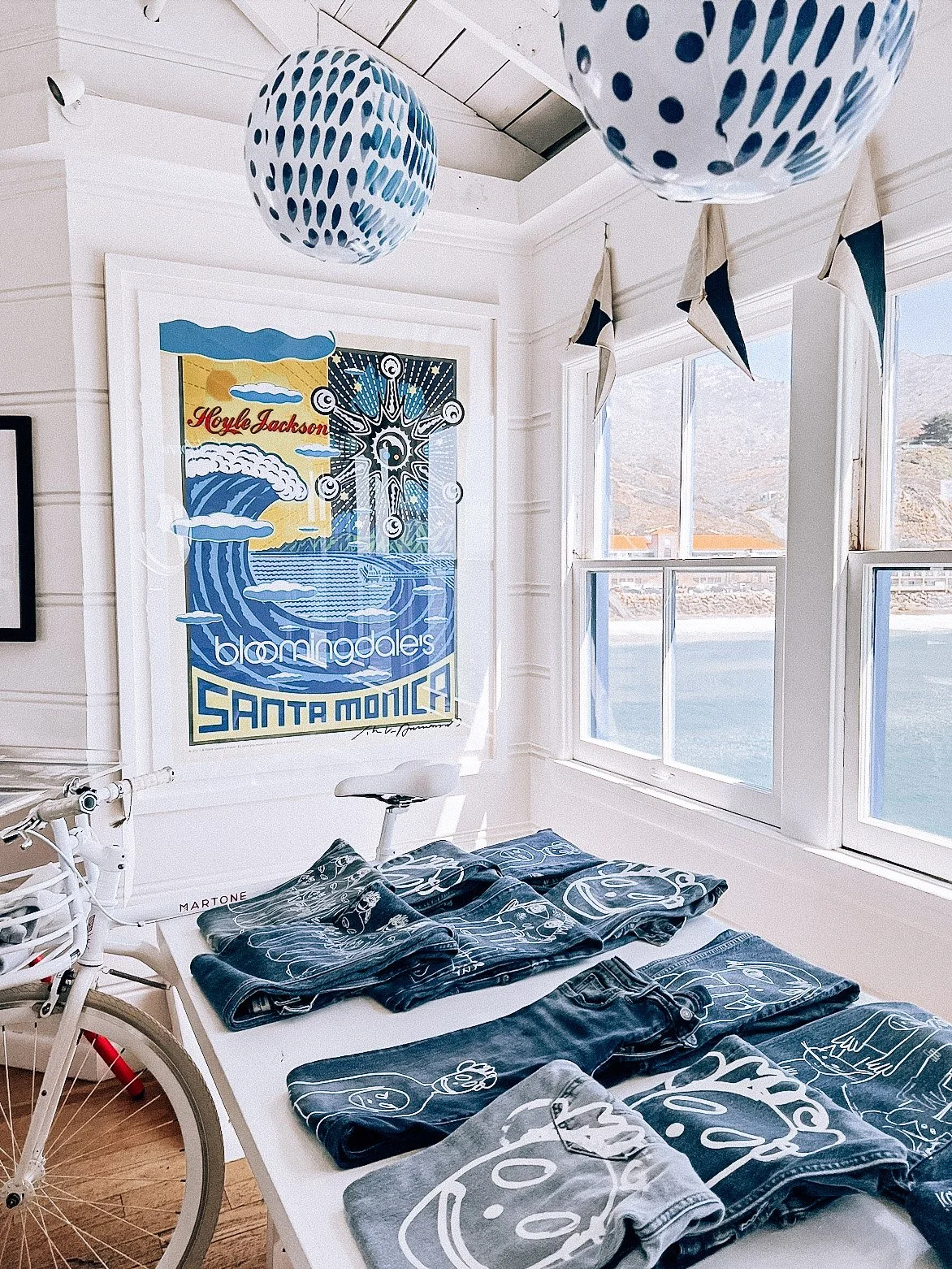 Room with white walls and large windows overlooking a body of water and hills. Decor includes blue and white paper lanterns hanging from the ceiling, a large framed poster on the wall for Bloomingdale's Santa Monica, and folded denim jeans and jacket