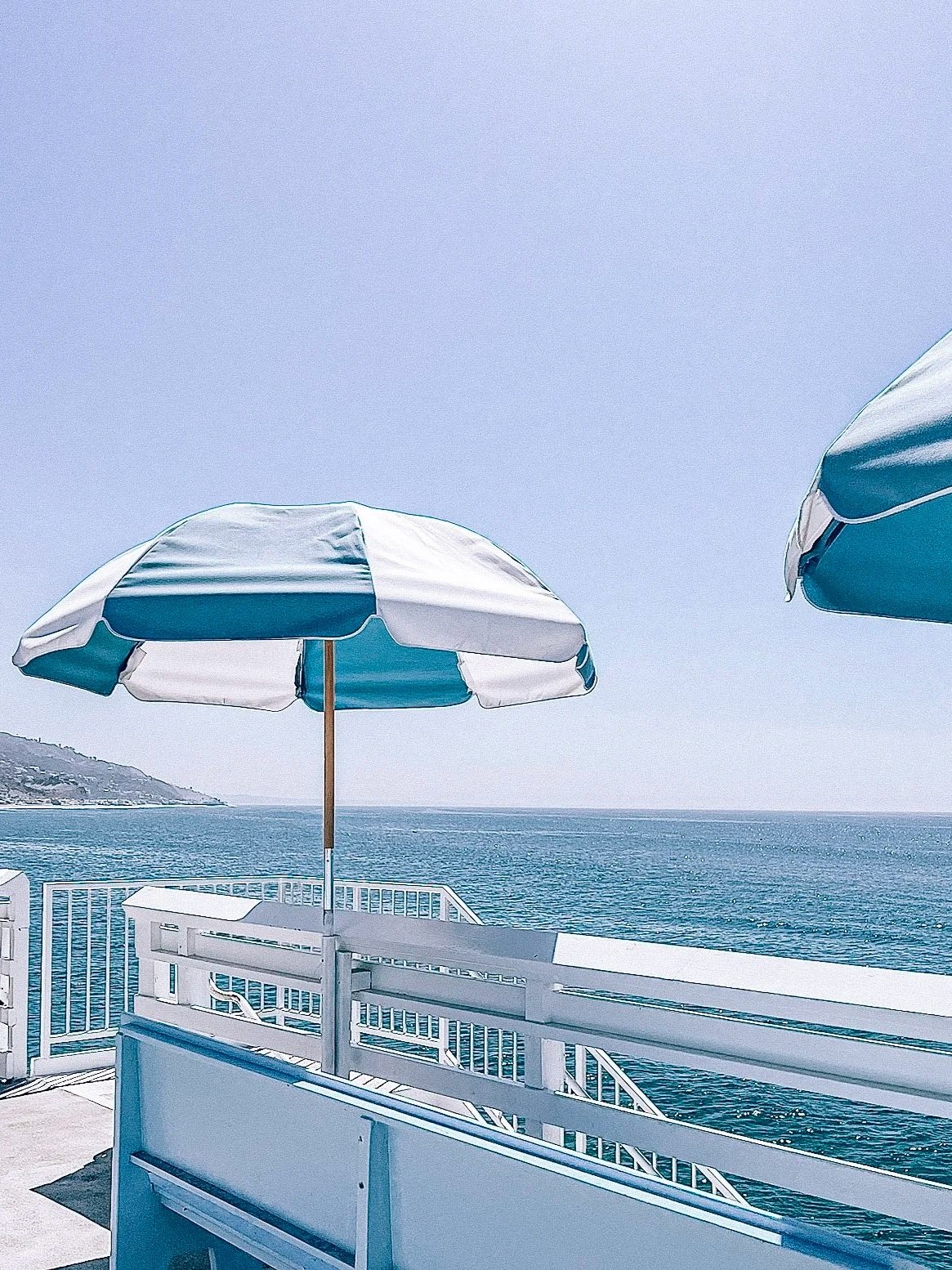 View of a beachside deck with a white and blue striped umbrella, calm ocean water in the background, and a distant hillside under a clear sky.