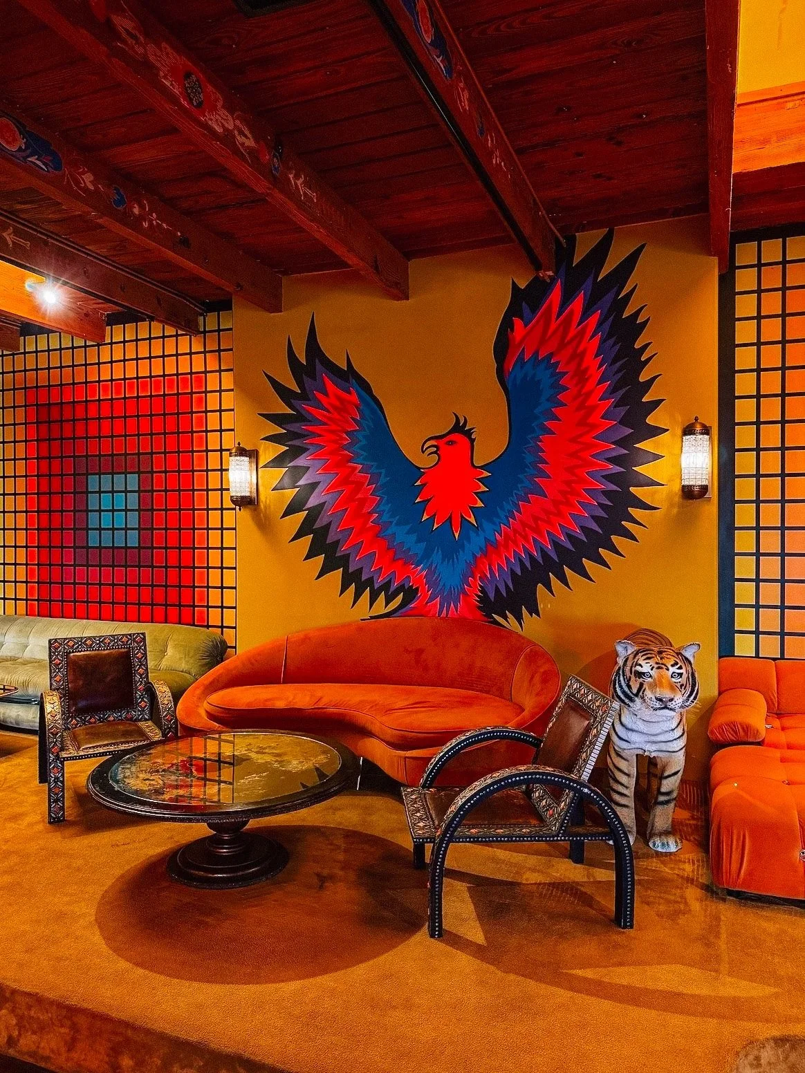 A colorful room with a large mural of a stylized red and blue phoenix on a yellow wall. The room has orange and green sofas, wicker chairs, and a glass-top wooden table. There is a stuffed tiger figure on one of the sofas, and the walls have a grid p