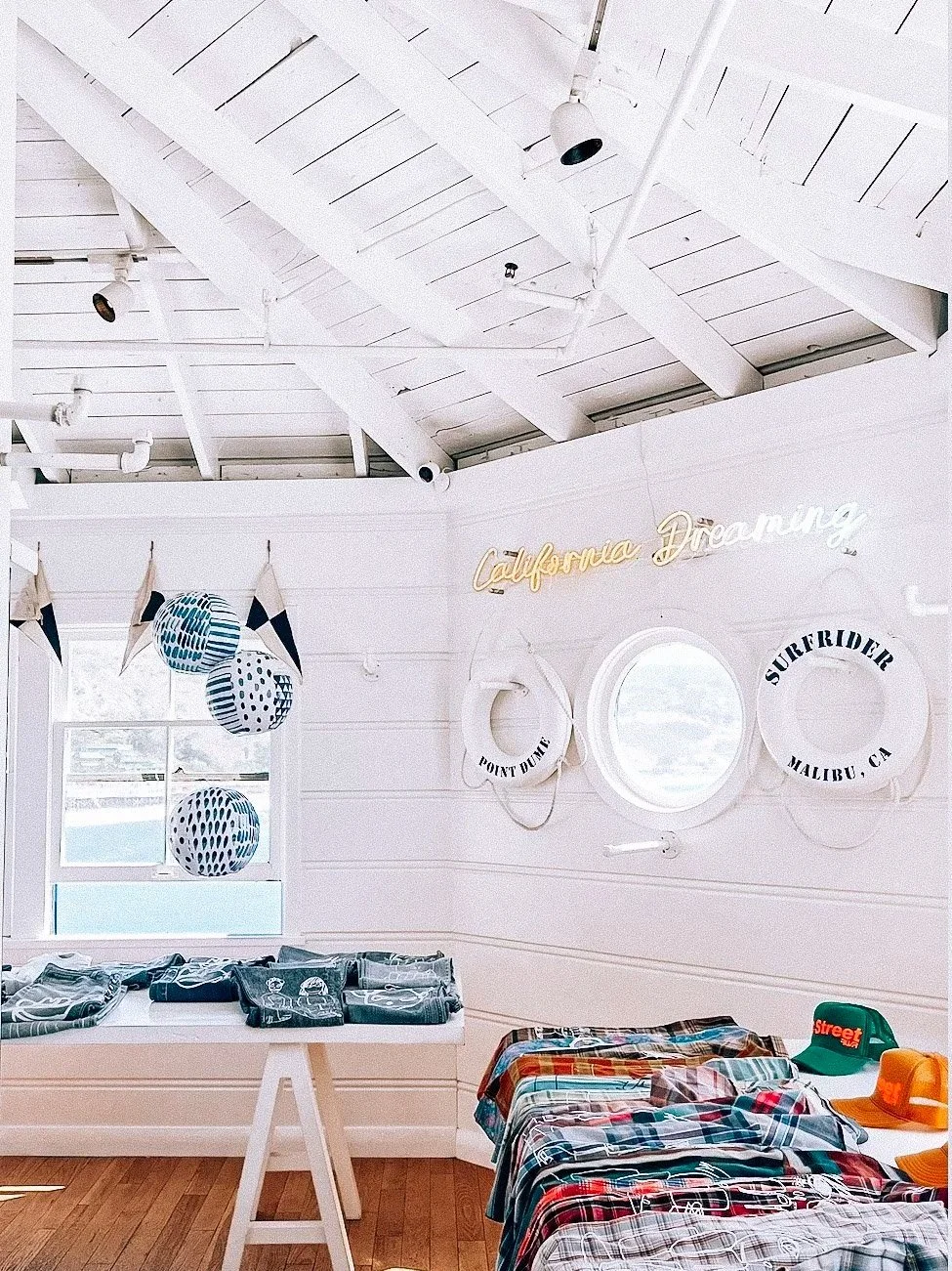 Interior of a beach-themed retail store with white wooden walls and ceiling, nautical decorations, and clothing displayed on tables.
