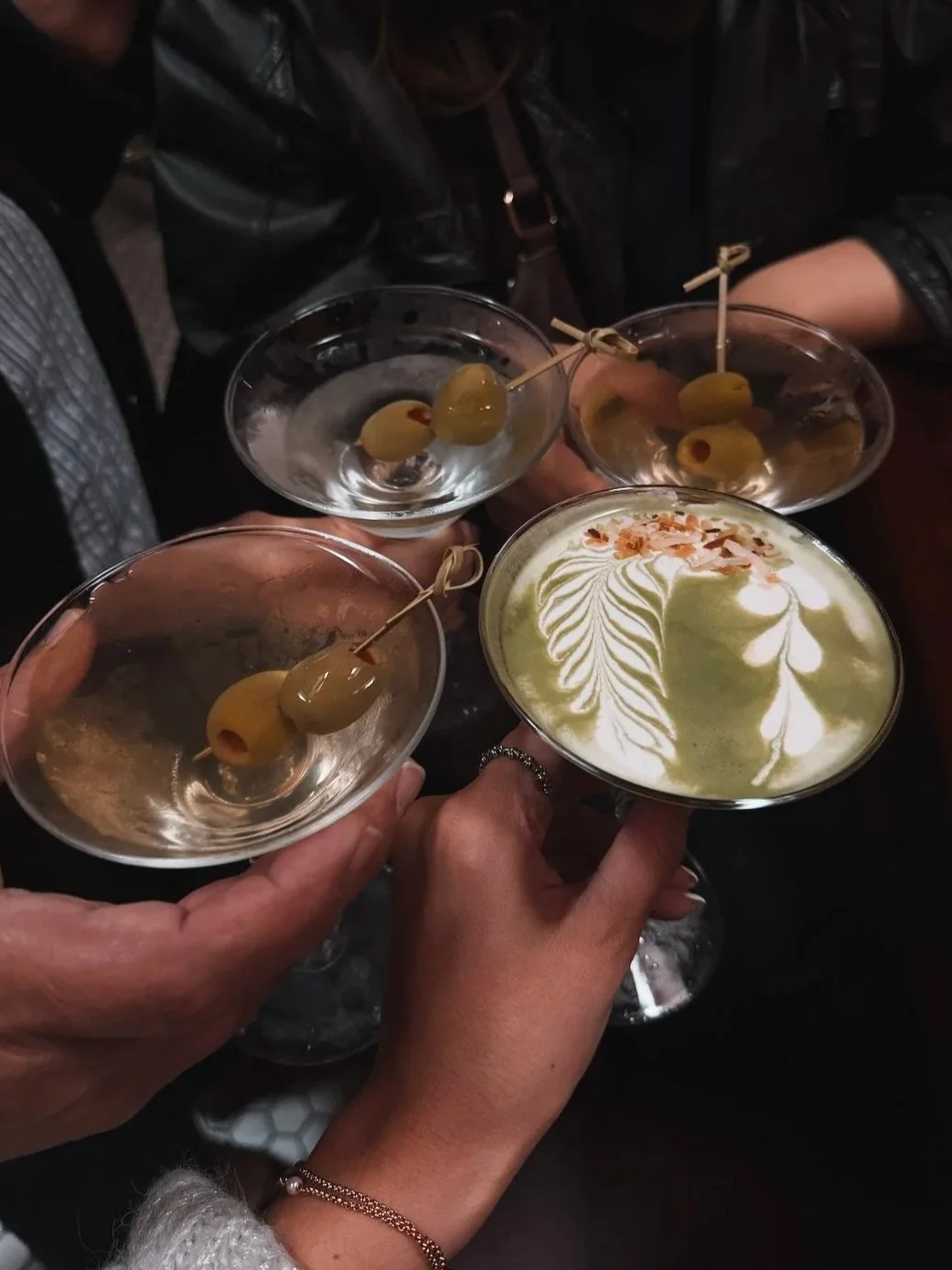 People holding cocktail glasses with green olives, and a drink with latte art on top.