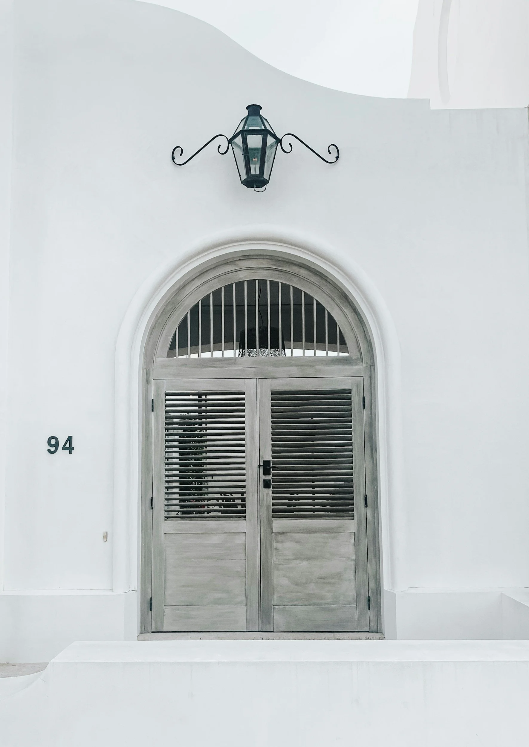 A white building wall with a grey double door featuring horizontal slats and a rounded arch window at the top. The number 94 is displayed to the left of the door, and a black decorative outdoor lantern is mounted above the door.