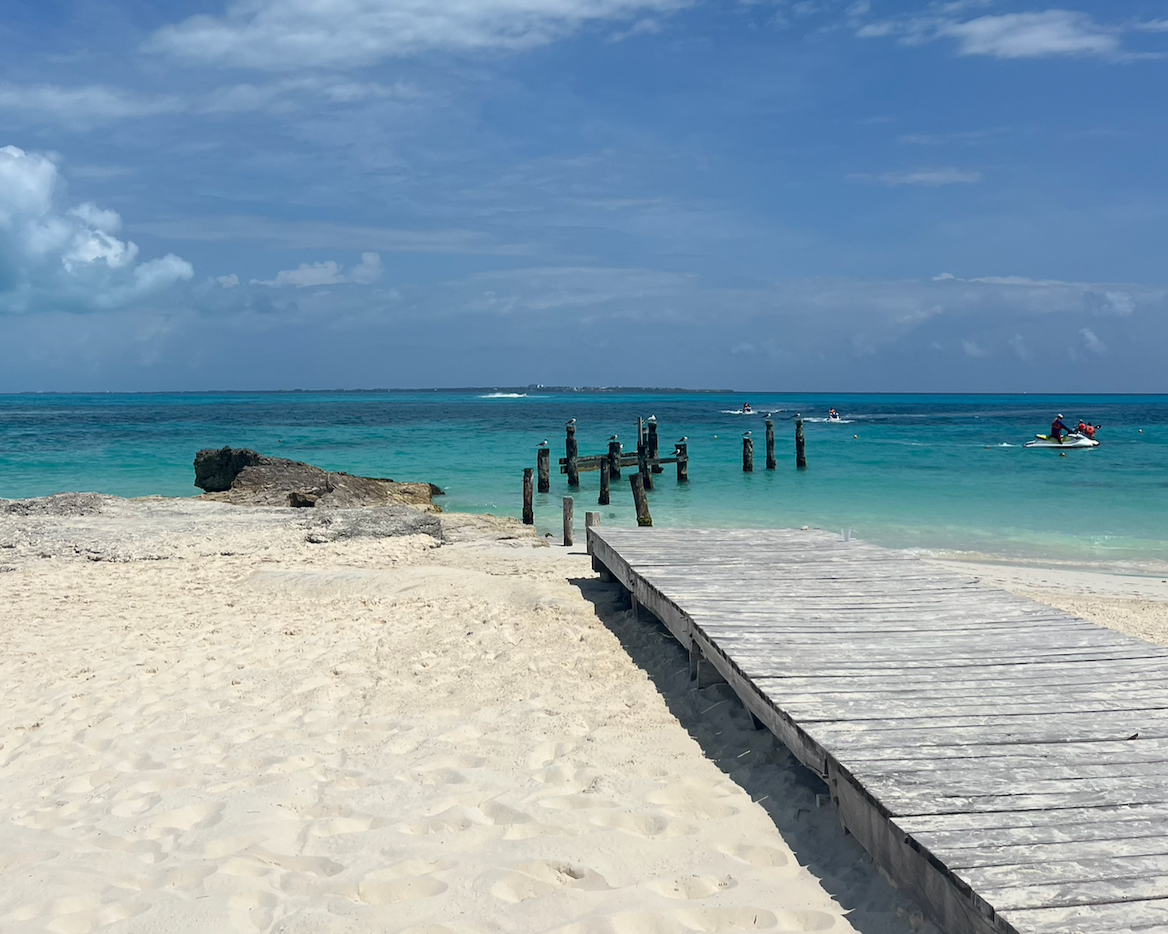 A sandy beach with a wooden dock leading into turquoise water, with pilings in the water and boats in the distance under a partly cloudy sky.