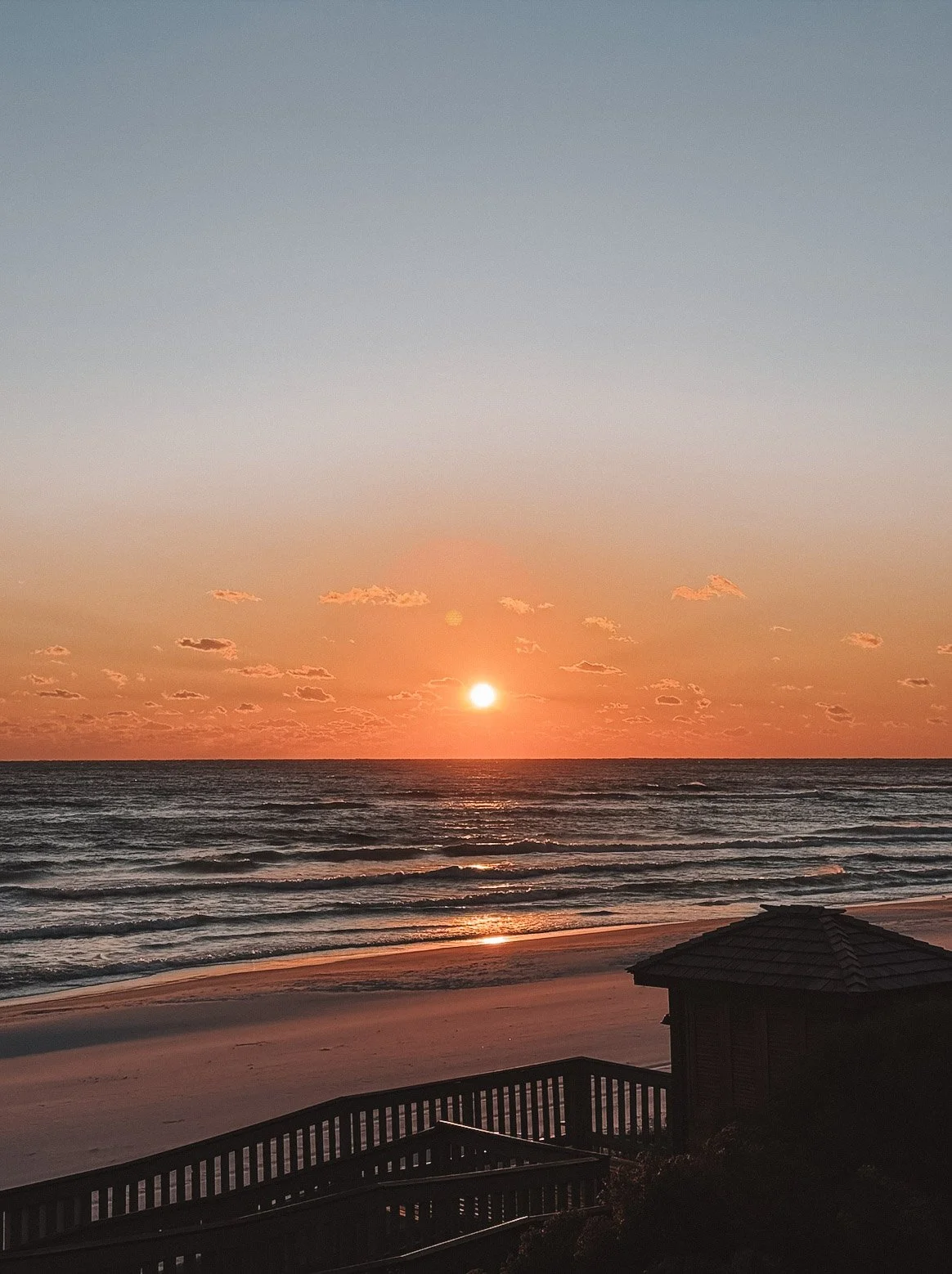 Sunset over the ocean with a partly cloudy sky and a wooden beach house in the foreground.