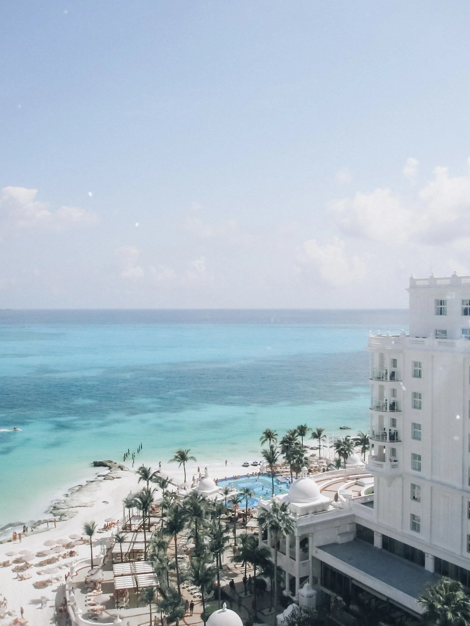 A beachside resort with a sandy beach, palm trees, a swimming pool, and a white multi-story hotel building overlooking the ocean, under a partly cloudy sky.