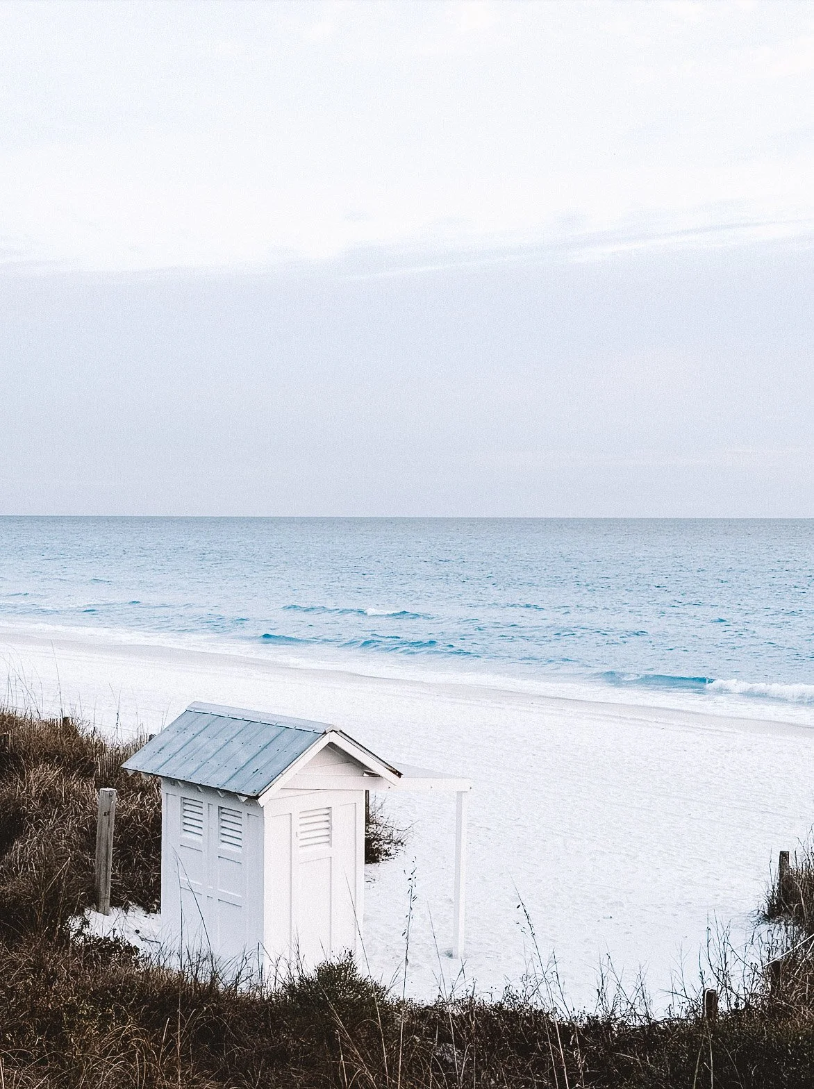 A small white beach hut with a metal roof, situated on sandy dunes with sparse grasses, overlooking the ocean with gentle waves under a cloudy sky.