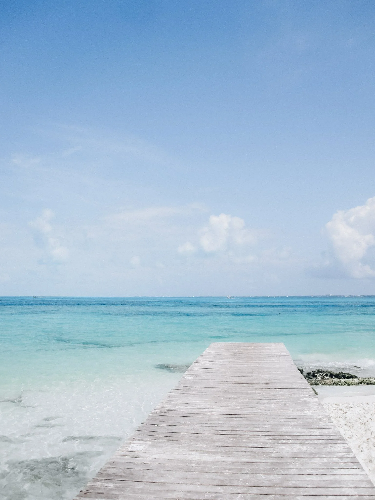 A wooden pier extends into the ocean with clear turquoise water, leading to a horizon with a blue sky and some white clouds.