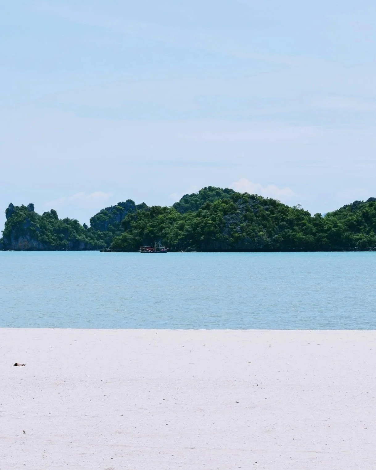 A tropical beach with white sand, calm blue water, and lush green islands in the distance under a partly cloudy sky.