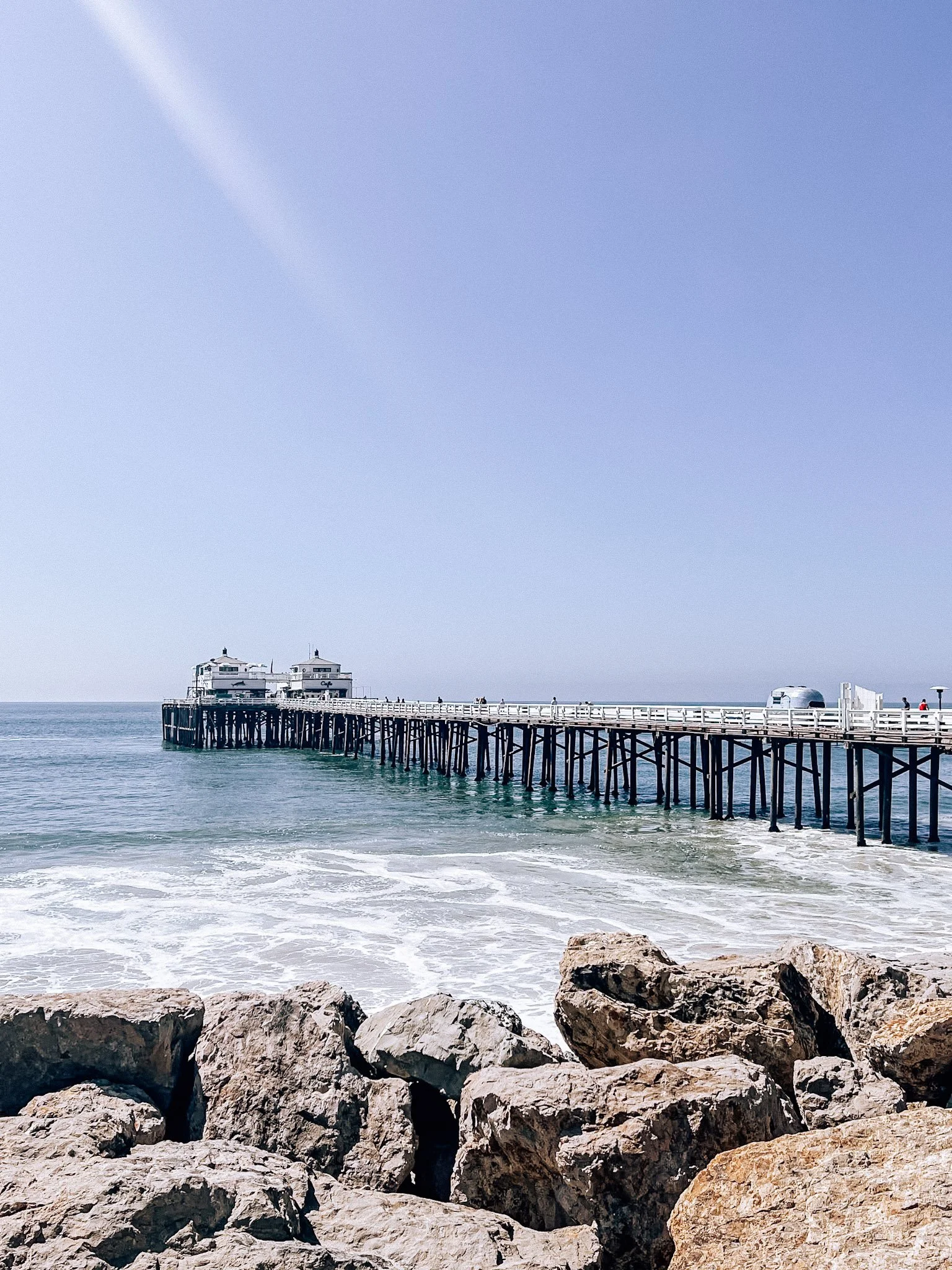 A pier extending into the ocean with rocks in the foreground, water, and a clear blue sky.
