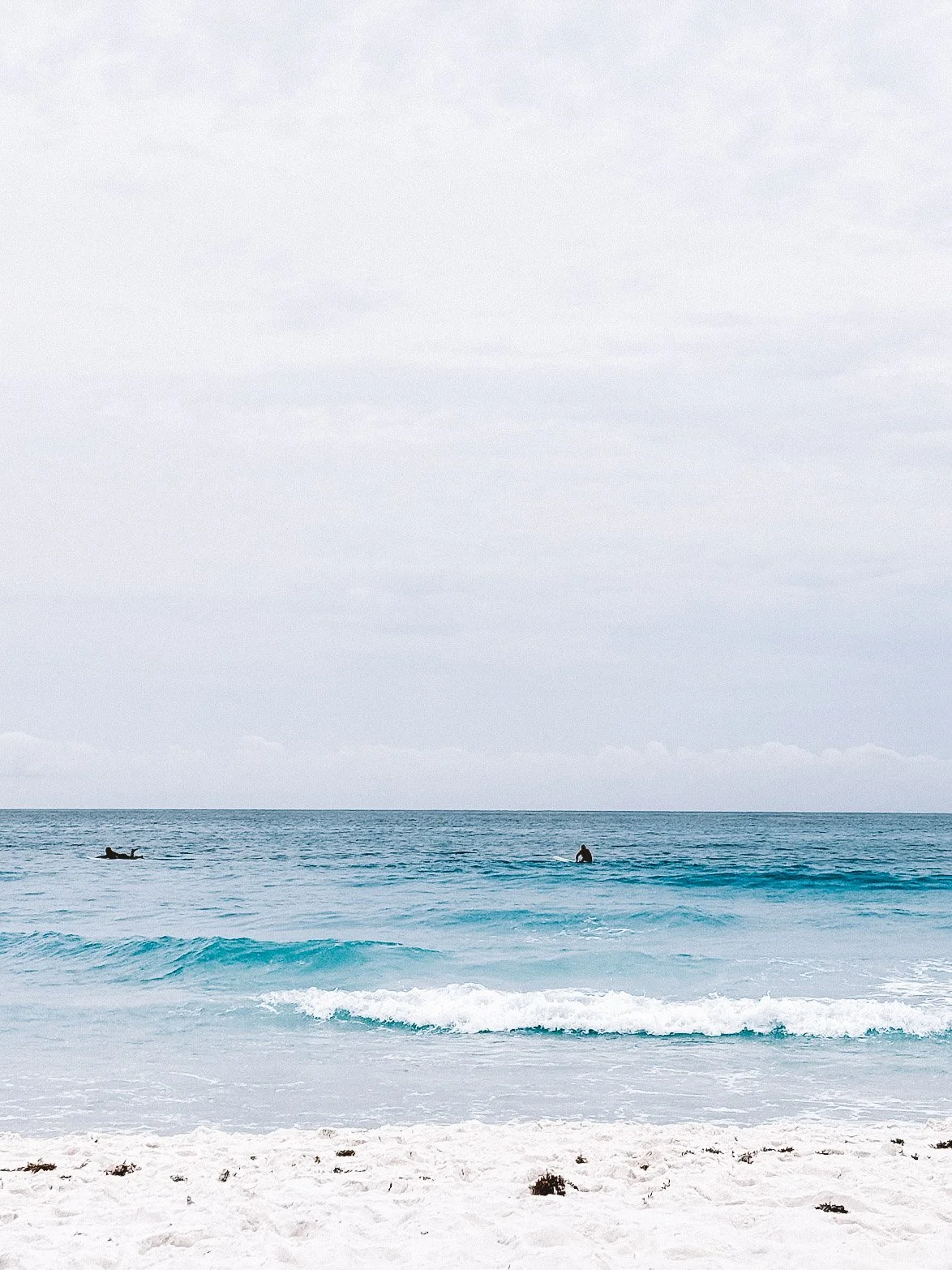 Calm ocean with waves washing onto sandy beach, two surfers in the distance, overcast sky.