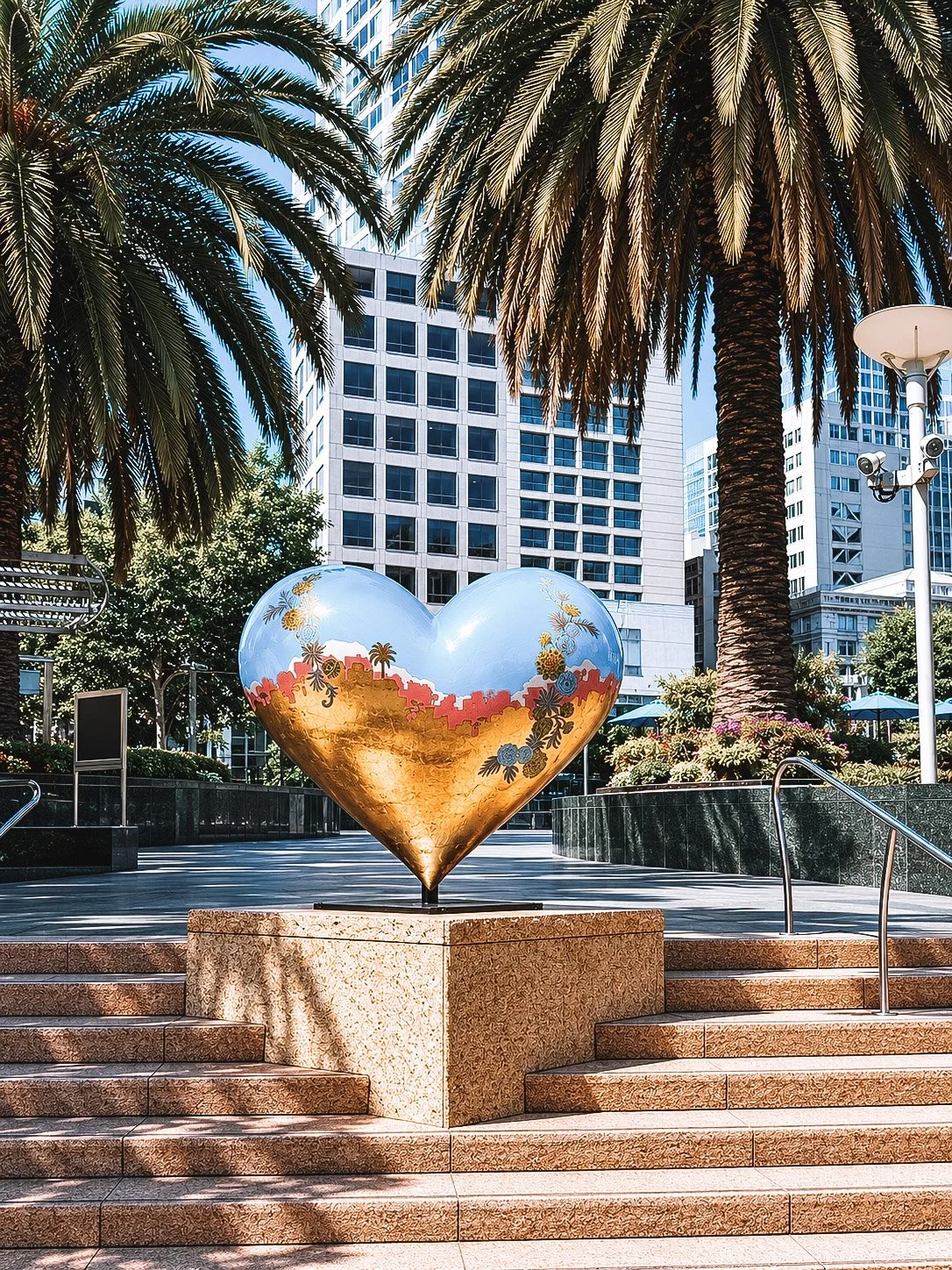 A large heart-shaped sculpture painted with a landscape scene, positioned on steps in an urban plaza surrounded by palm trees, modern office buildings, and decorative planters with flowers.