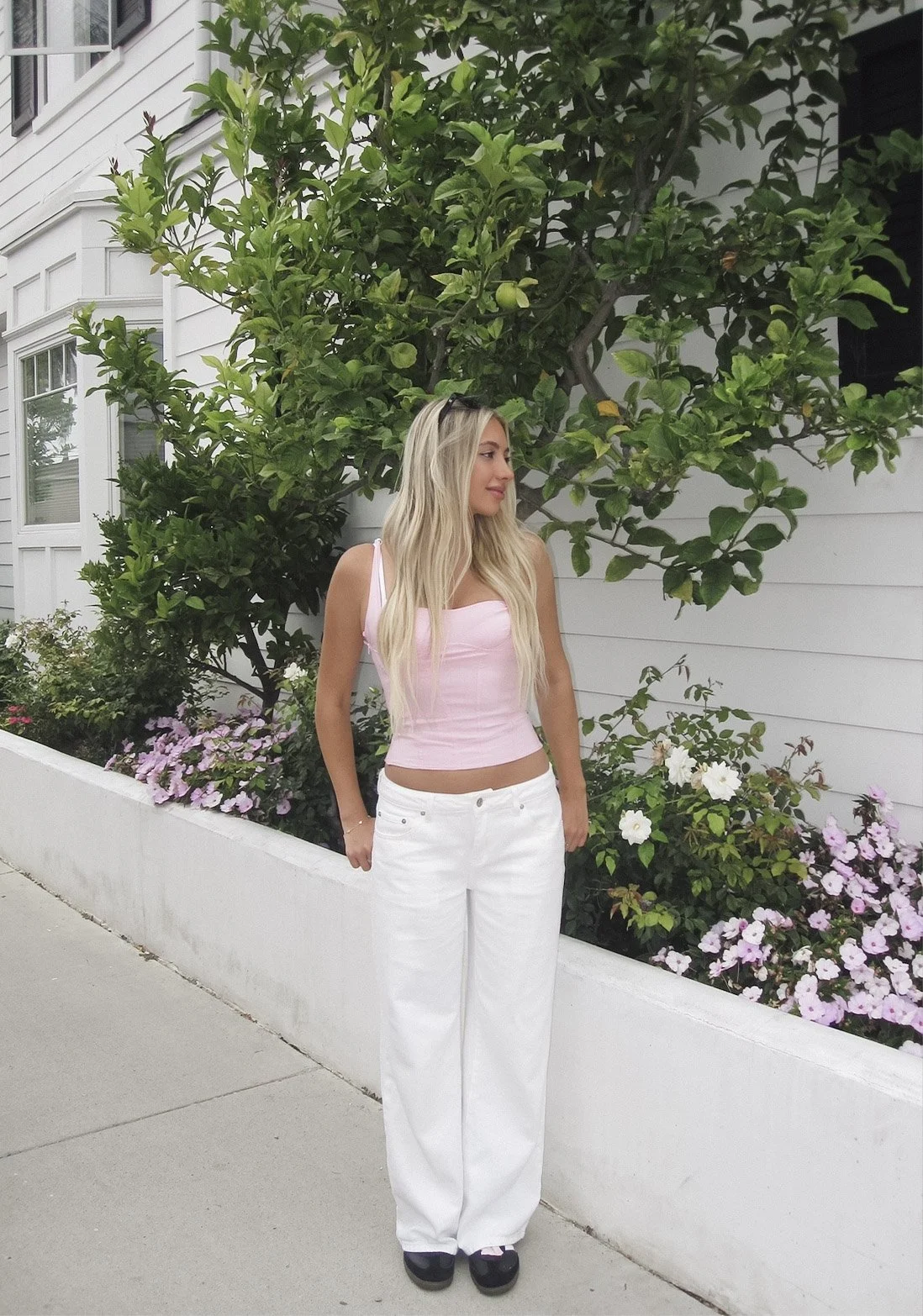 A woman in a pink sleeveless top and white pants standing next to a flowerbed with various pink and white flowers, in front of a white wall and greenery.