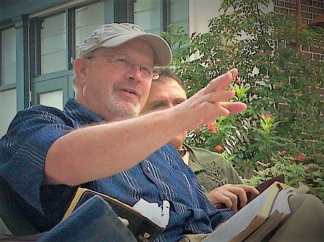 Two men sitting outdoors, one wearing glasses and a hat, gesturing with his hand, surrounded by greenery and a building in the background.