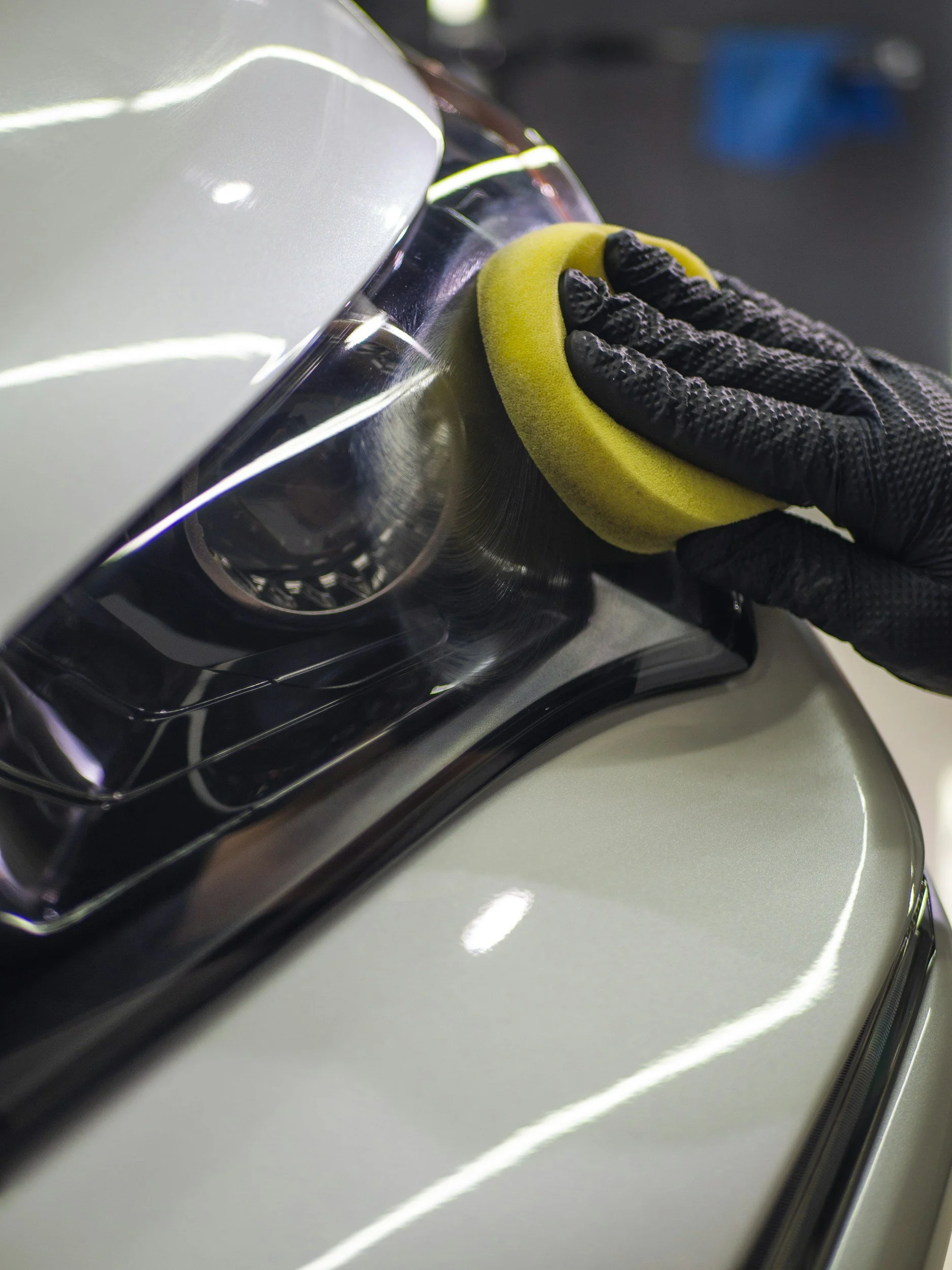 A person wearing a black glove polishing a silver car with a yellow polishing pad.