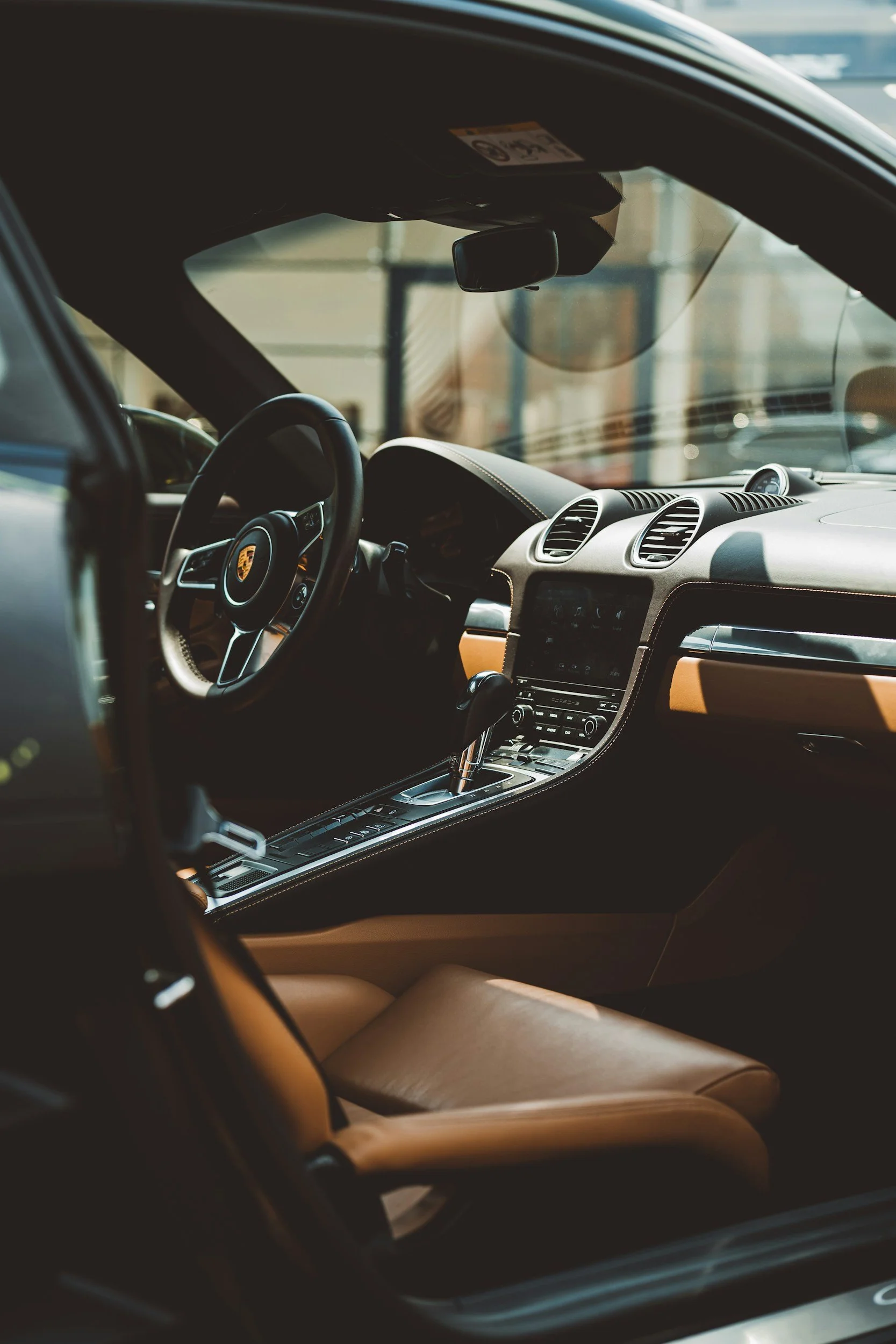 Inside view of a luxury car's interior showing the steering wheel with the Porsche logo in the center, center console with controls, and tan leather seats.