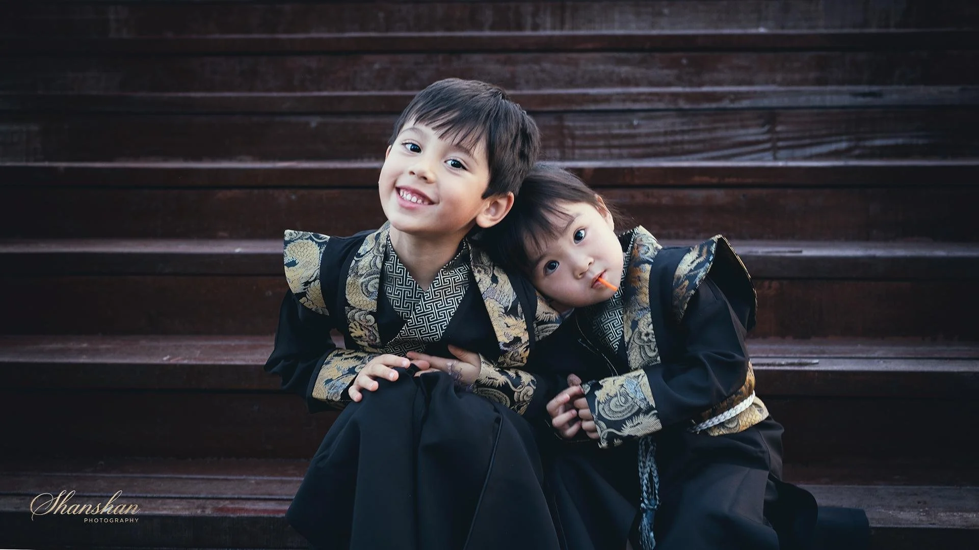 Two young boys wearing black and gold patterned traditional clothing sitting on wooden stairs, one smiling at the camera with head resting on the other's shoulder, the younger with a lollipop in mouth.