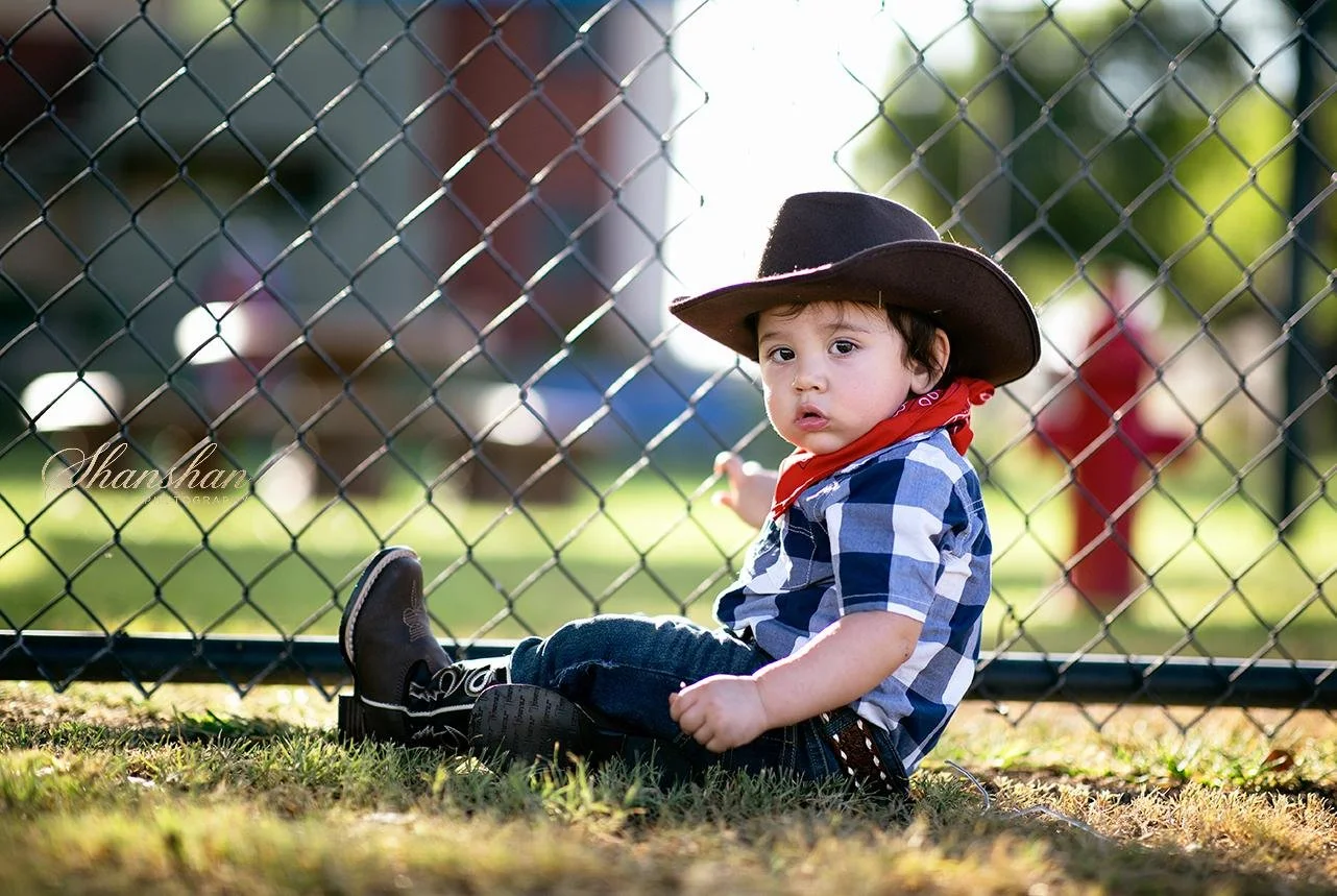 A young boy dressed as a cowboy, sitting on the grass near a chain-link fence, wearing a cowboy hat, red neckerchief, plaid shirt, jeans, and cowboy boots, with a blurred playground in the background.