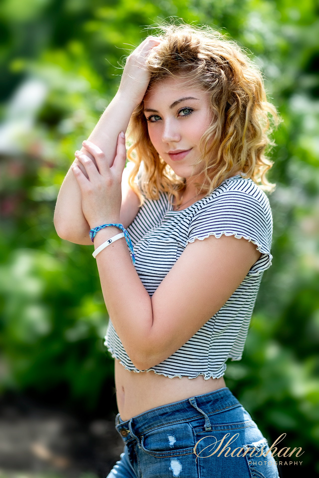 A young woman with curly blonde hair, wearing a striped crop top and ripped jeans, standing outdoors with greenery in the background, looking at the camera and touching her hair.