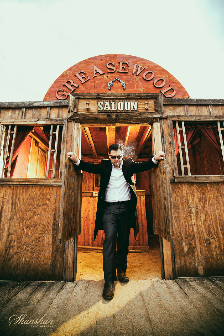 A man in a black coat and sunglasses walking out of a wooden saloon with a sign reading 'Greaswood' and a gun illustration above it.