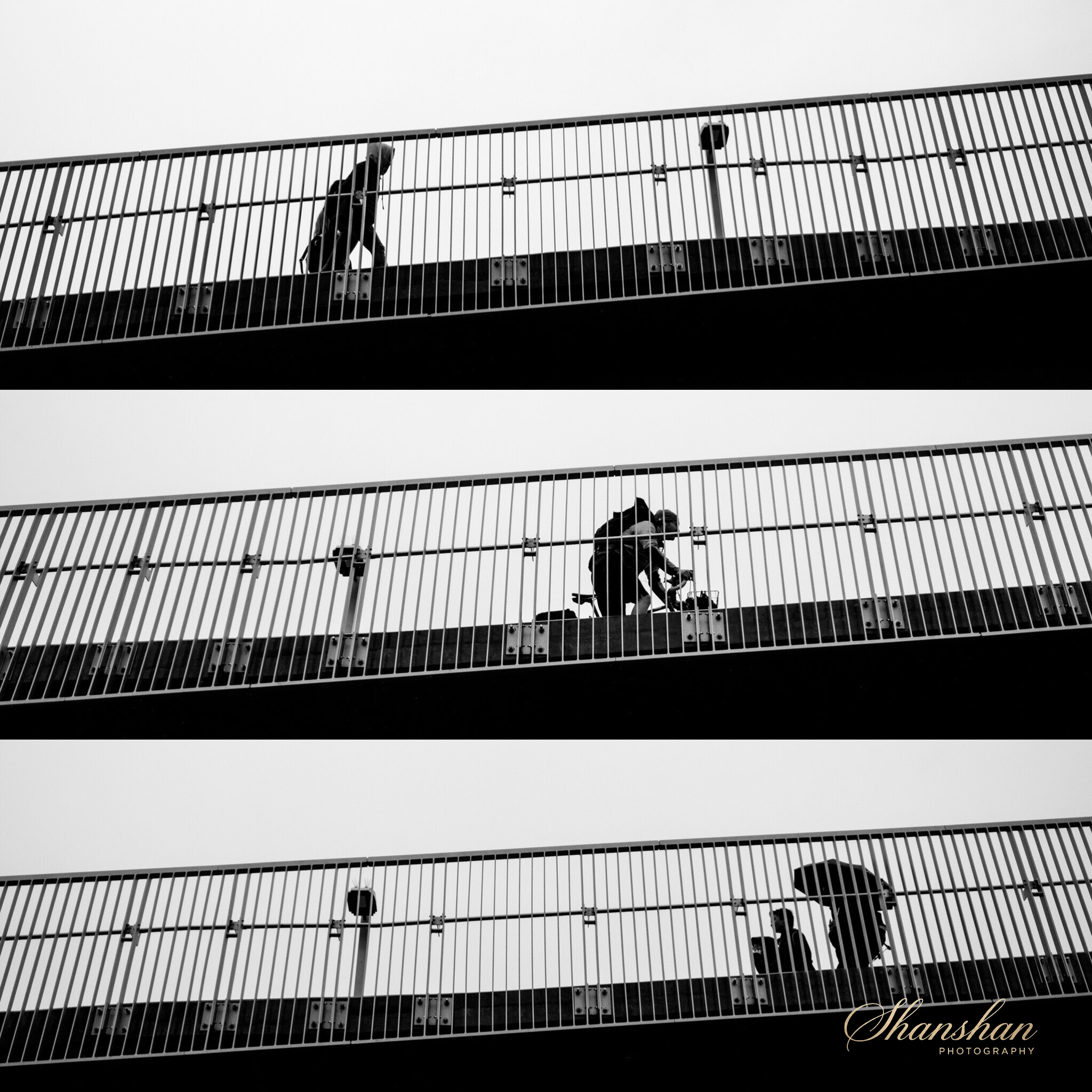A person dressed in black, wearing a hoodie, walking on a modern elevated pedestrian bridge with metal railings and support beams, captured in black and white.
