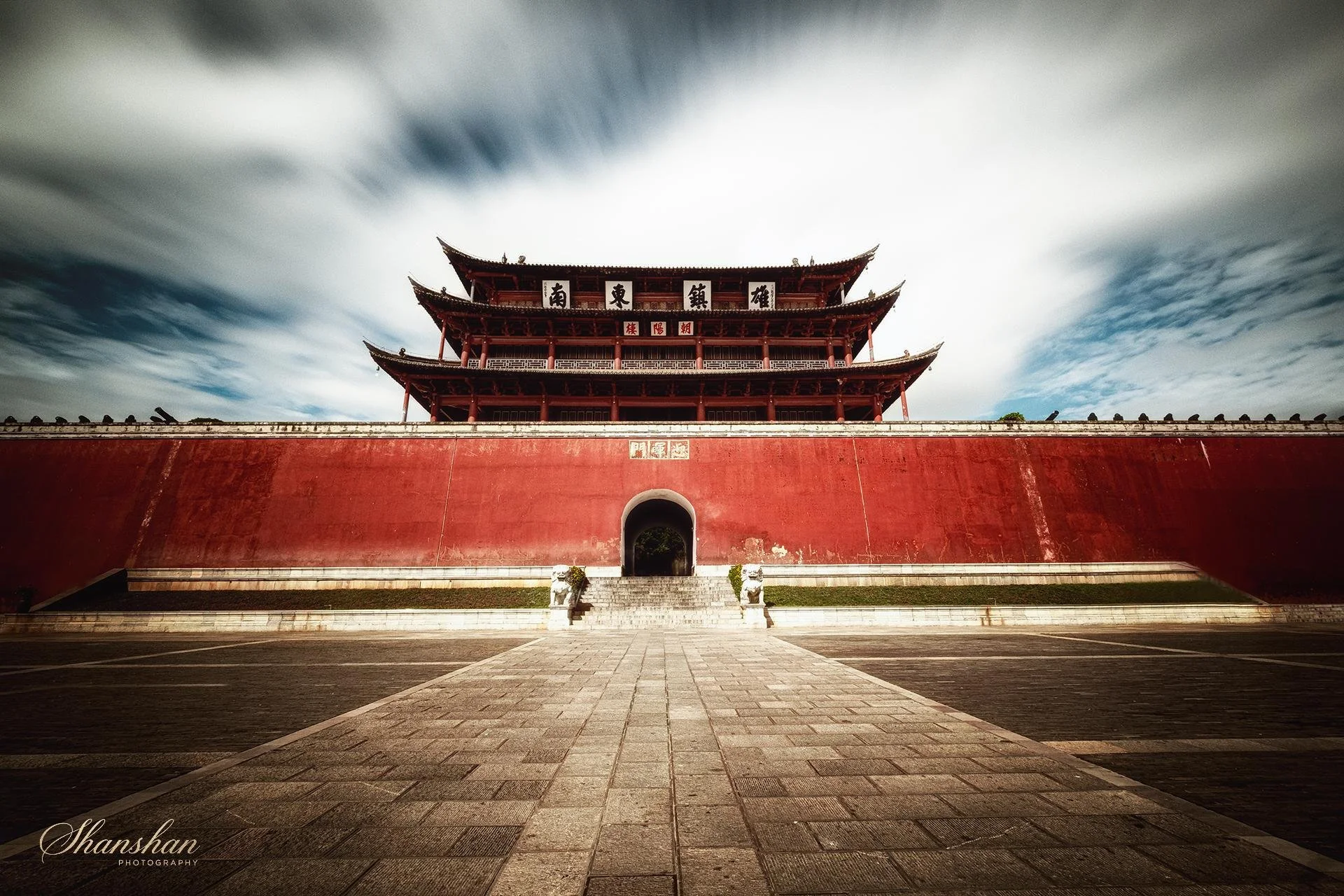 Ancient Chinese building with multiple tiers, red walls, and pagoda-style roof against a cloudy sky.
