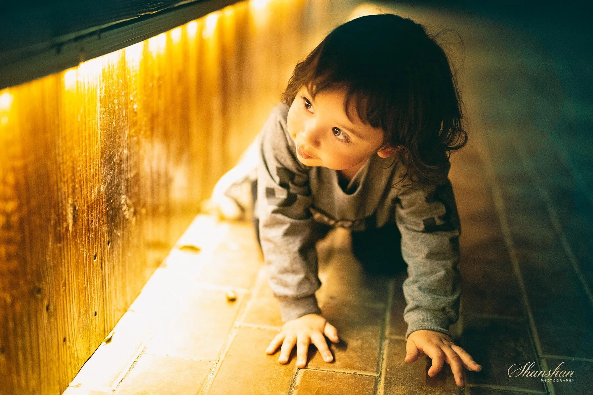 Young boy crawling on a tiled floor in an indoor setting with a warm, yellow lighting. He has curly dark hair and is wearing a gray sweatshirt with patterned sleeves.