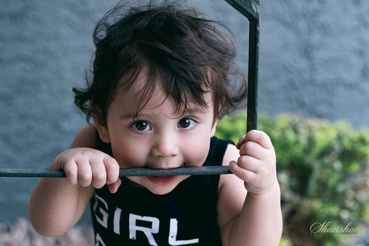 Young child with curly dark hair and big eyes, playfully holding onto a metal bar while peeking through it outdoors