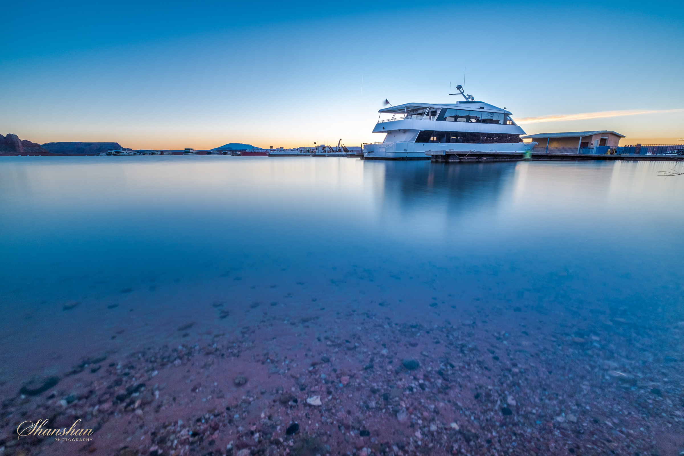A docked boat on a calm body of water during sunset or sunrise, with a smooth water surface and a clear sky in the background.
