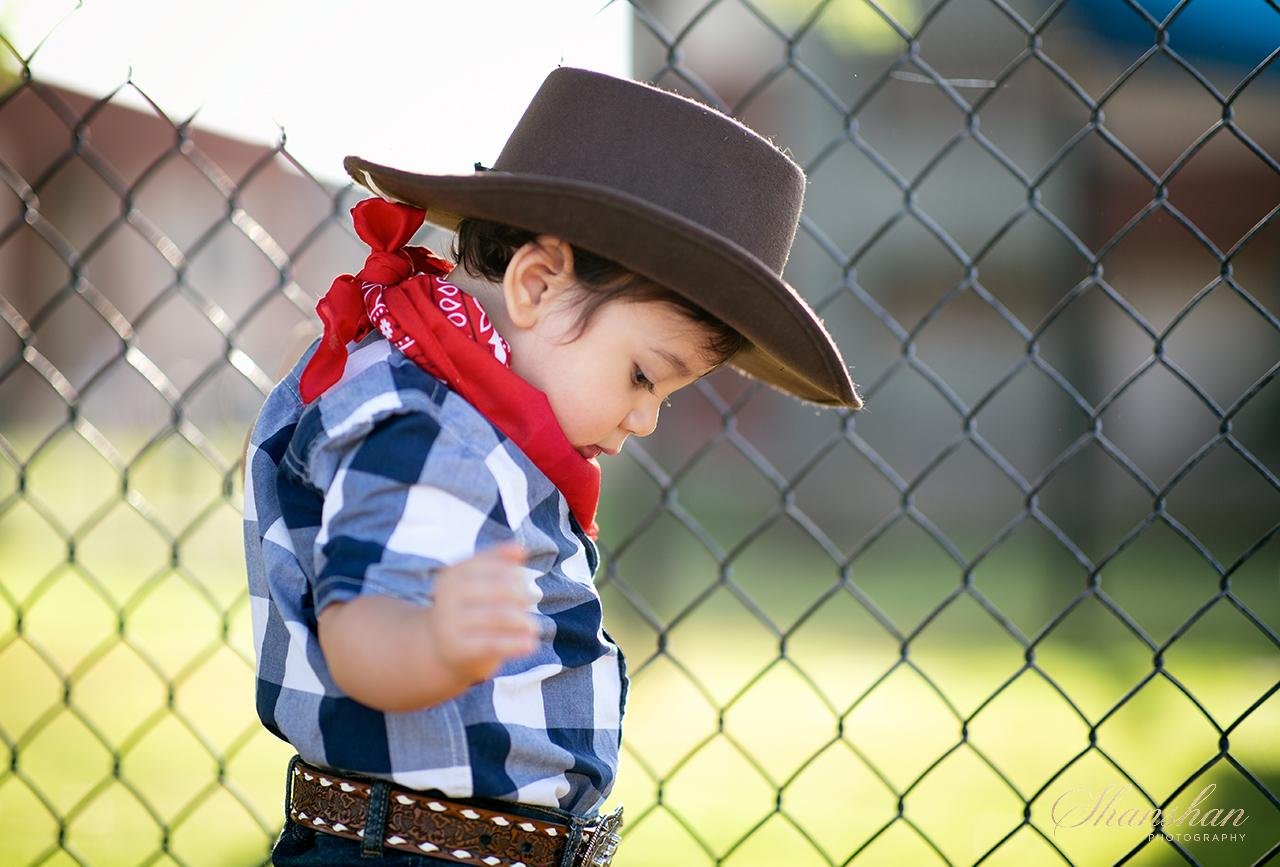 A young boy dressed as a cowboy, wearing a brown cowboy hat, a red bandana, and a blue checkered shirt, standing next to a chain-link fence with a blurry outdoor background.