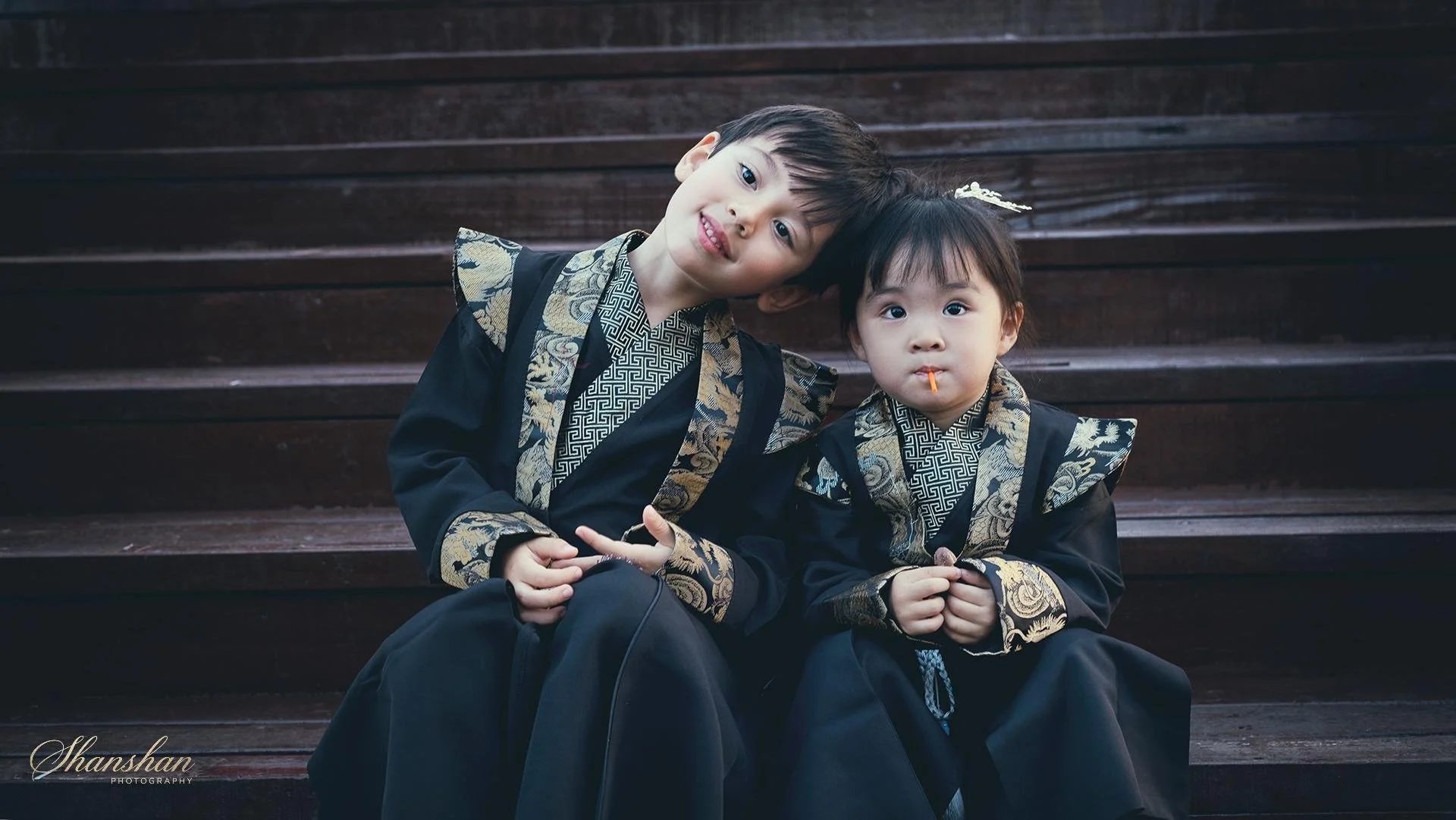 Two children dressed in traditional black outfits with gold and silver embroidery, sitting on wooden steps. The older boy is leaning his head on the younger girl, who has a lollipop in her mouth. They are outdoors, and the background shows dark woode