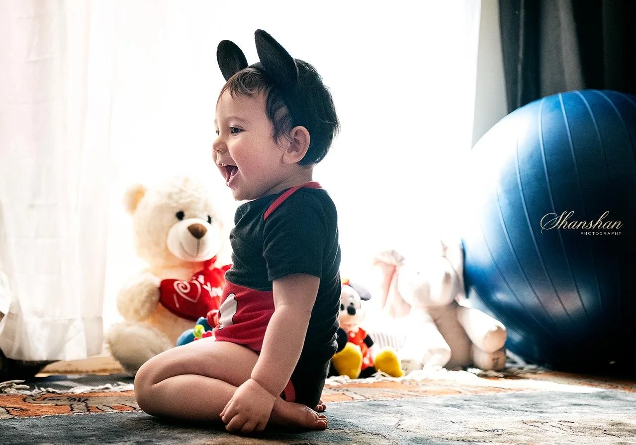 A young child sitting on a carpeted floor, wearing a black and red outfit with black animal ears headband, surrounded by plush toys including a teddy bear, a dog, and an elephant, in front of a bright window.