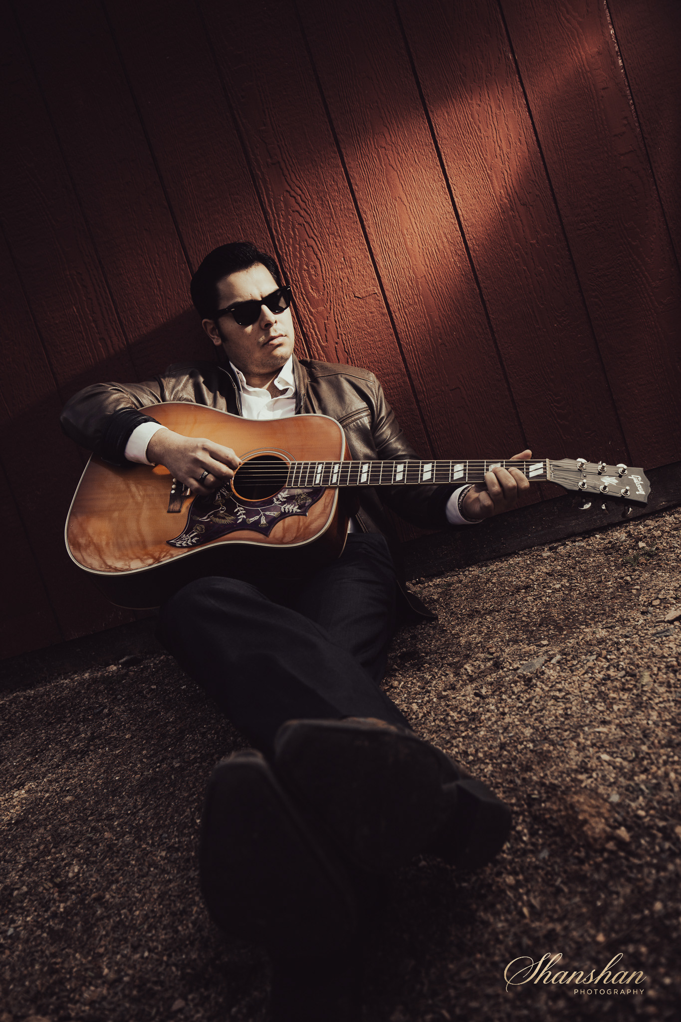 A man sitting against a wooden wall playing an acoustic guitar, wearing sunglasses and a leather jacket, with a serious expression.