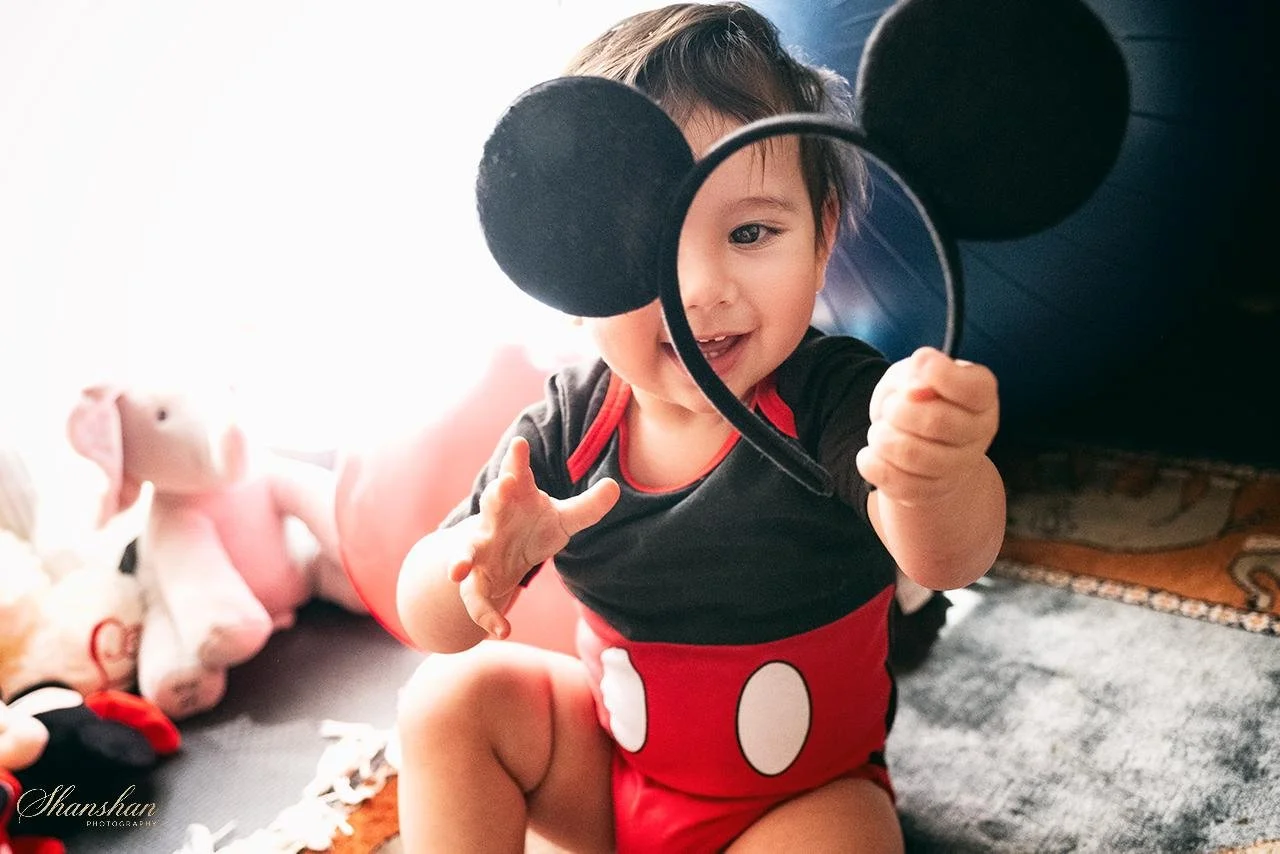 A young child dressed in Mickey Mouse pajamas, holding a mirror, with Mickey Mouse ears on top, sitting on a carpeted floor surrounded by stuffed animals.