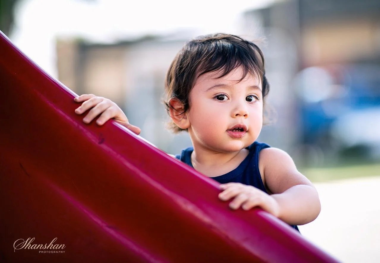 Young child with dark hair holding a red slide outdoors, wearing a dark sleeveless shirt, looking towards the camera with a curious expression.