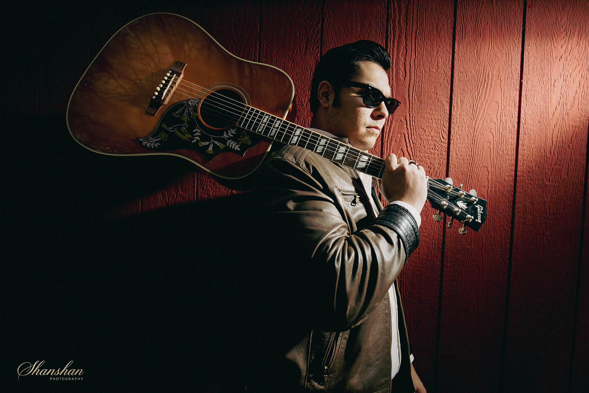 A man wearing sunglasses and a brown leather jacket is leaning against a red wooden wall, holding an acoustic guitar over his shoulder. He has dark, slicked-back hair and appears to be looking to the side.