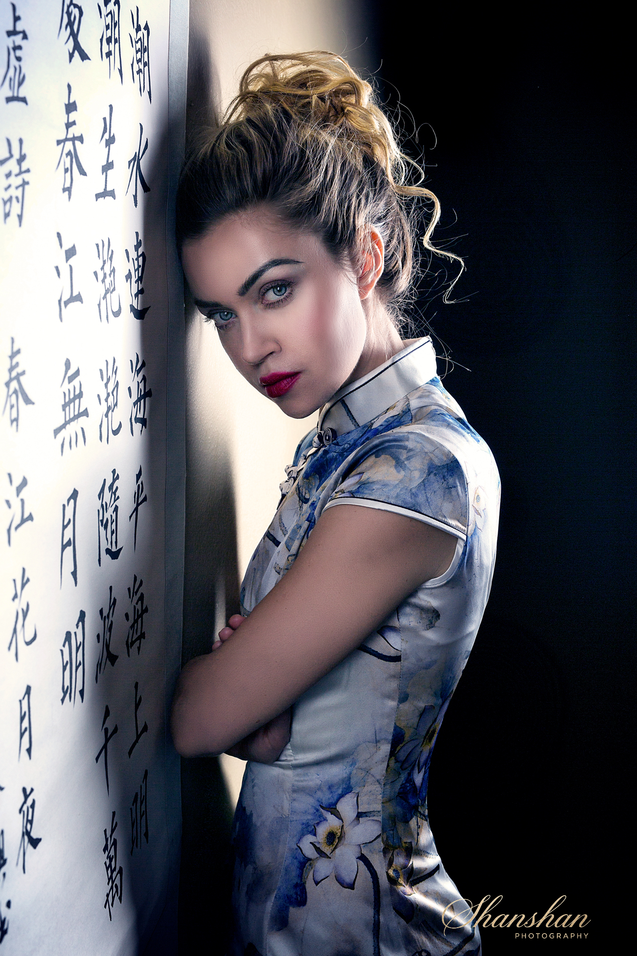 A woman with blue eyes, red lipstick, and styled hair, wearing a floral cheongsam, standing against a wall with Chinese characters, looking at the camera.