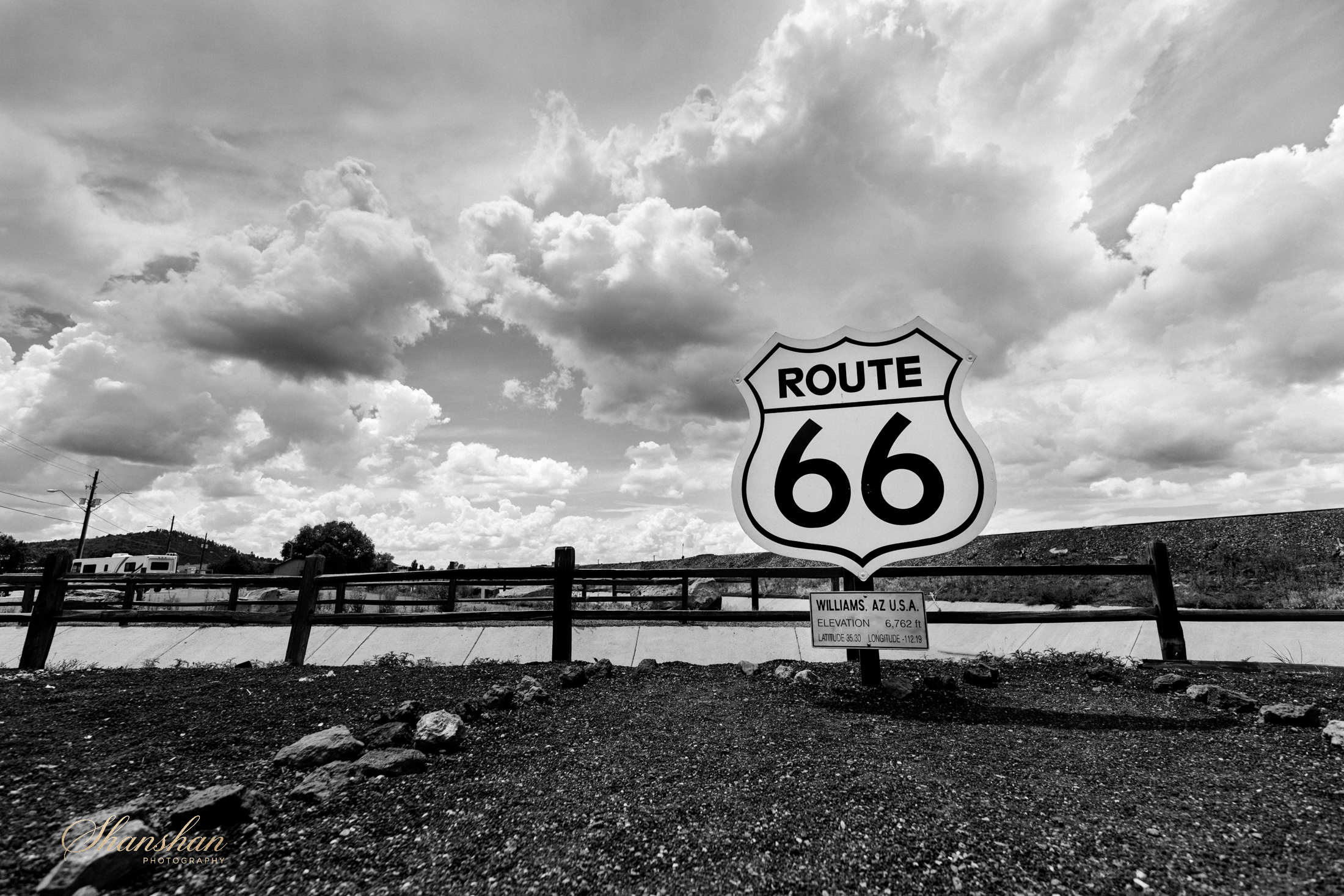 Black and white photo of a road sign for Route 66 with a partly cloudy sky and power lines in the background.
