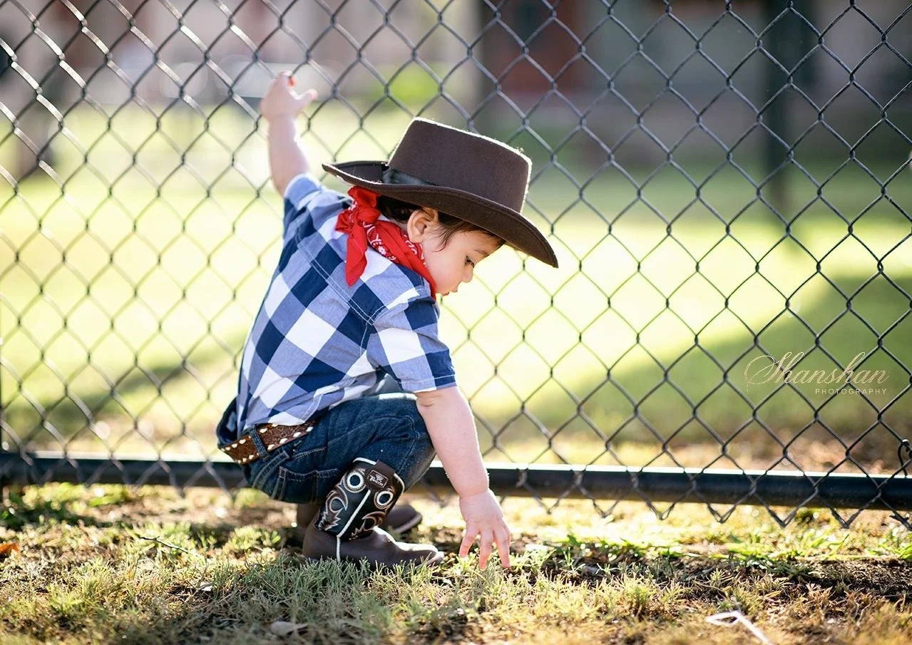 Young child dressed as a cowboy, wearing a large brown cowboy hat, red bandana, plaid shirt, jeans, and cowboy boots, crouching near a chain-link fence and touching grass on the ground.