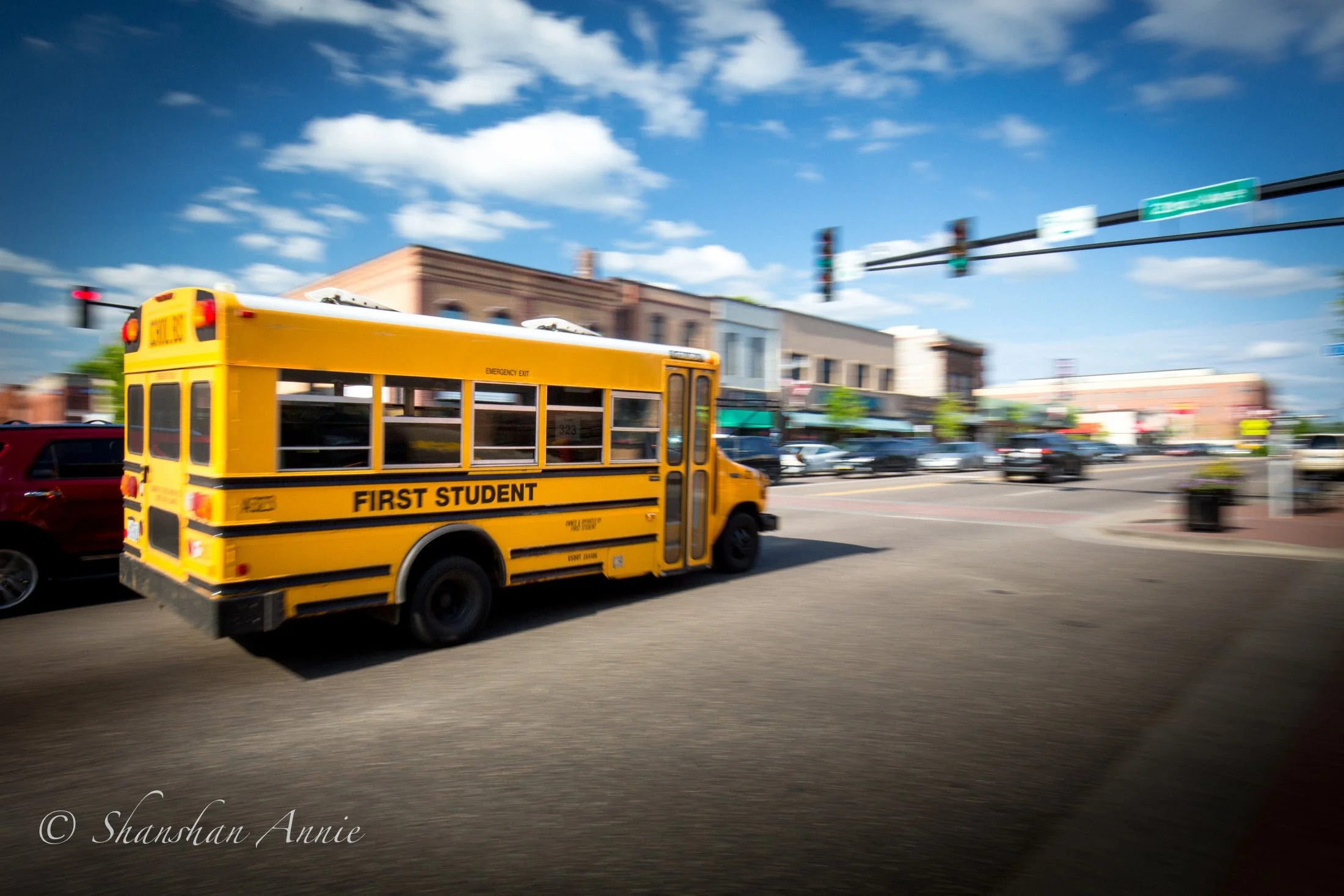 Yellow school bus with 'FIRST STUDENT' on the side driving through a small town on a sunny day, with buildings, parking lot, and traffic lights in the background.