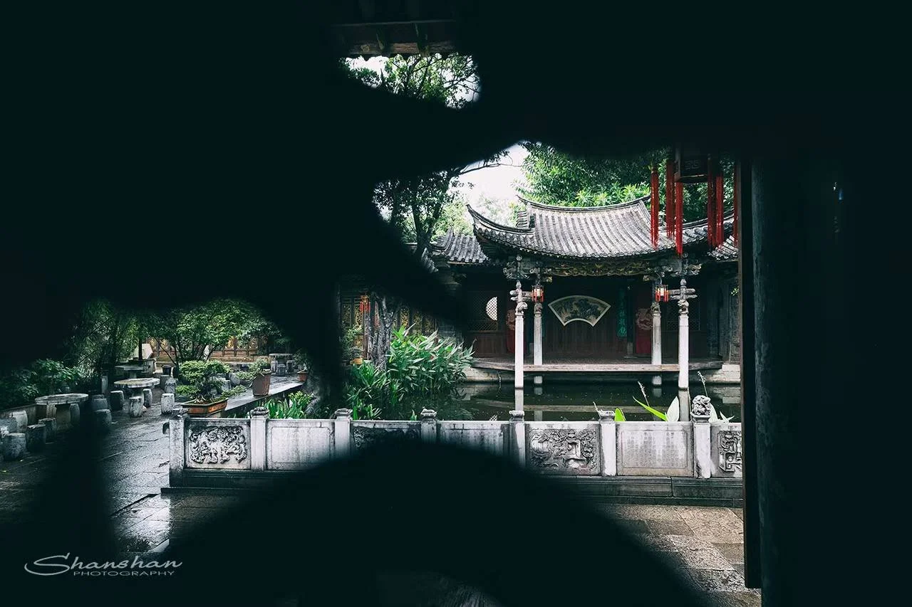 Traditional Chinese garden with a pavilion, stone pathway, greenery, and stone sculptures, seen through a dark opening.
