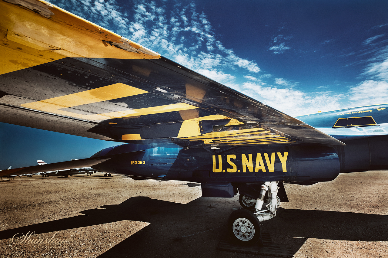 Close-up of a U.S. Navy fighter jet parked on a tarmac, showing the underside of the aircraft with blue and yellow markings, under a blue sky with scattered clouds.
