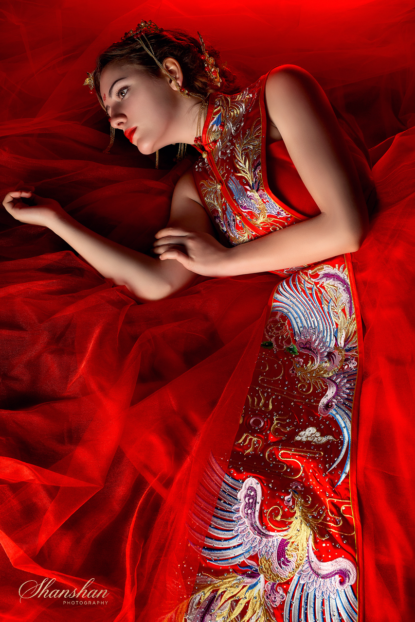 A woman dressed in a traditional red Chinese wedding dress with elaborate embroidery, lying on red tulle fabric, wearing ornate jewelry and hair accessories.