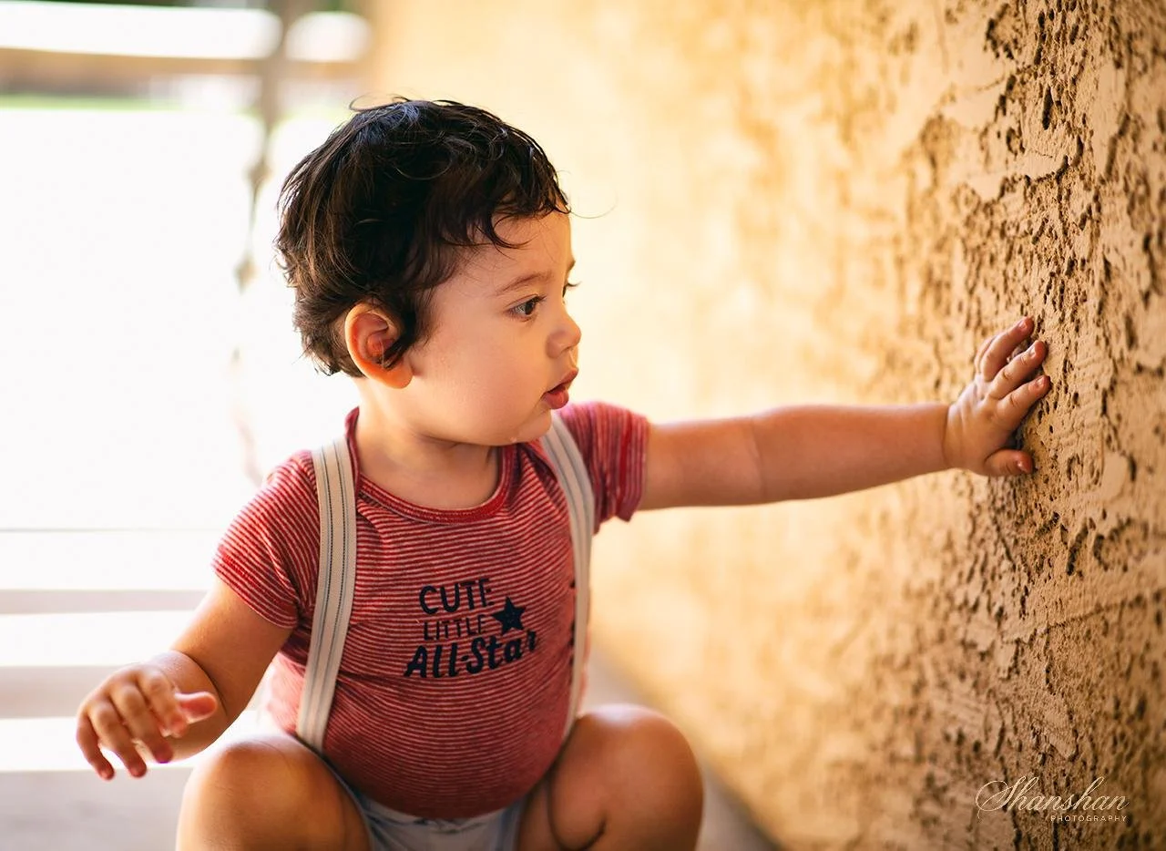 Young child wearing red striped shirt and suspenders, touching textured yellow wall while kneeling.