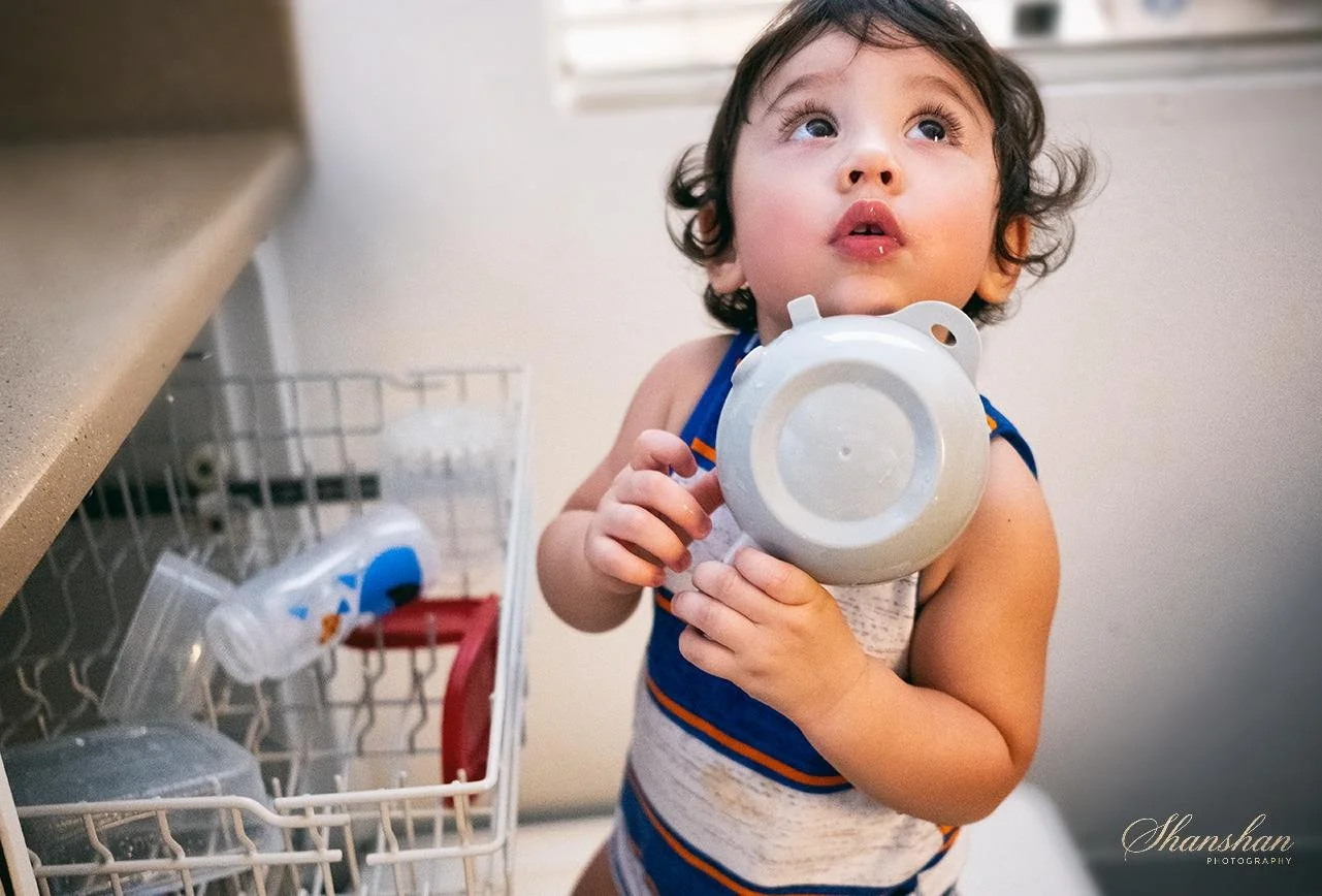 A young child with curly hair wearing a sleeveless striped shirt, holding a plastic plate near their mouth and looking up with a curious expression, standing next to a dishwasher in a kitchen.